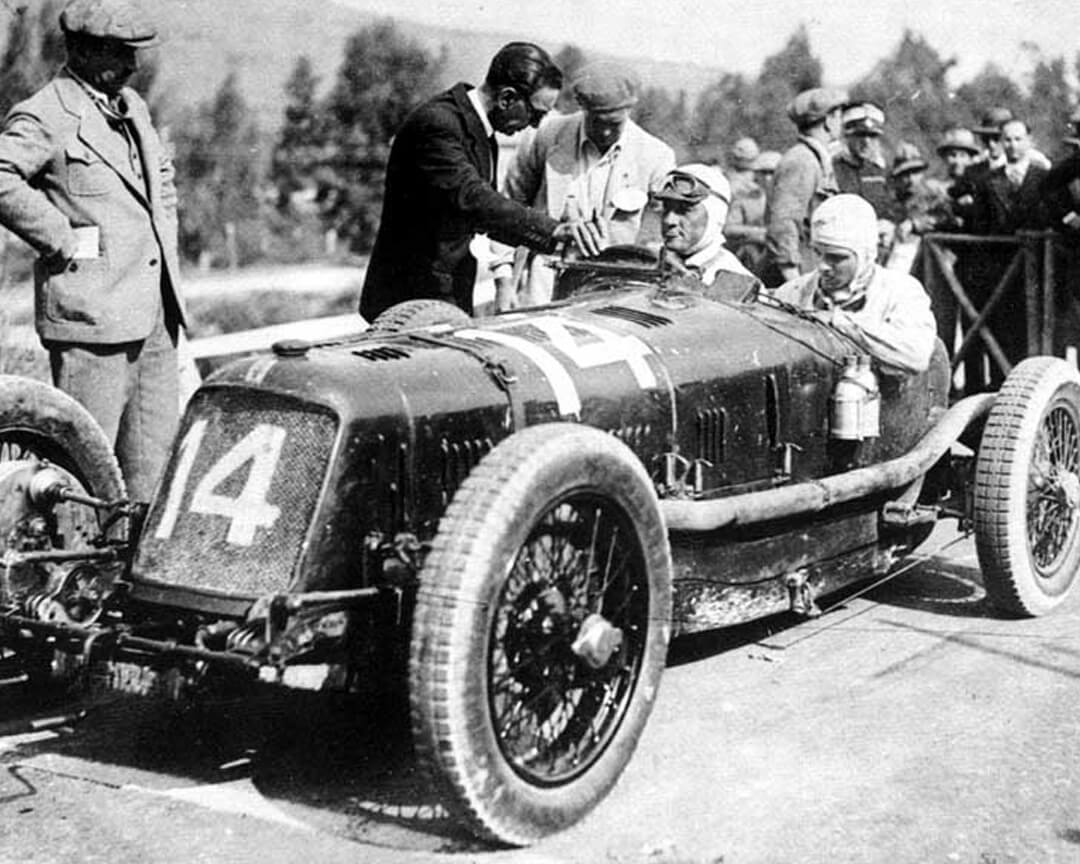 Foto d'epoca in bianco e nero di una vecchia maserati da corsa con il numero 14 e i due piloti alla guida pronti a partire