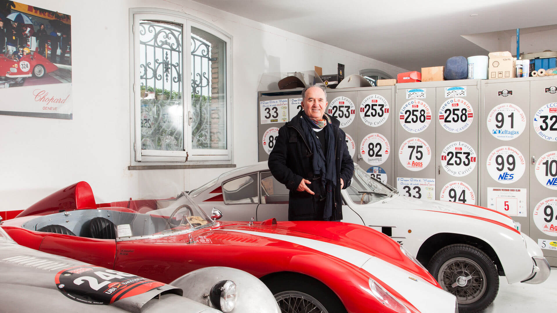 Foto di Ilario Bandini tra le sue auto d'epoca con alle spalle numeri appartenenti alle macchine
