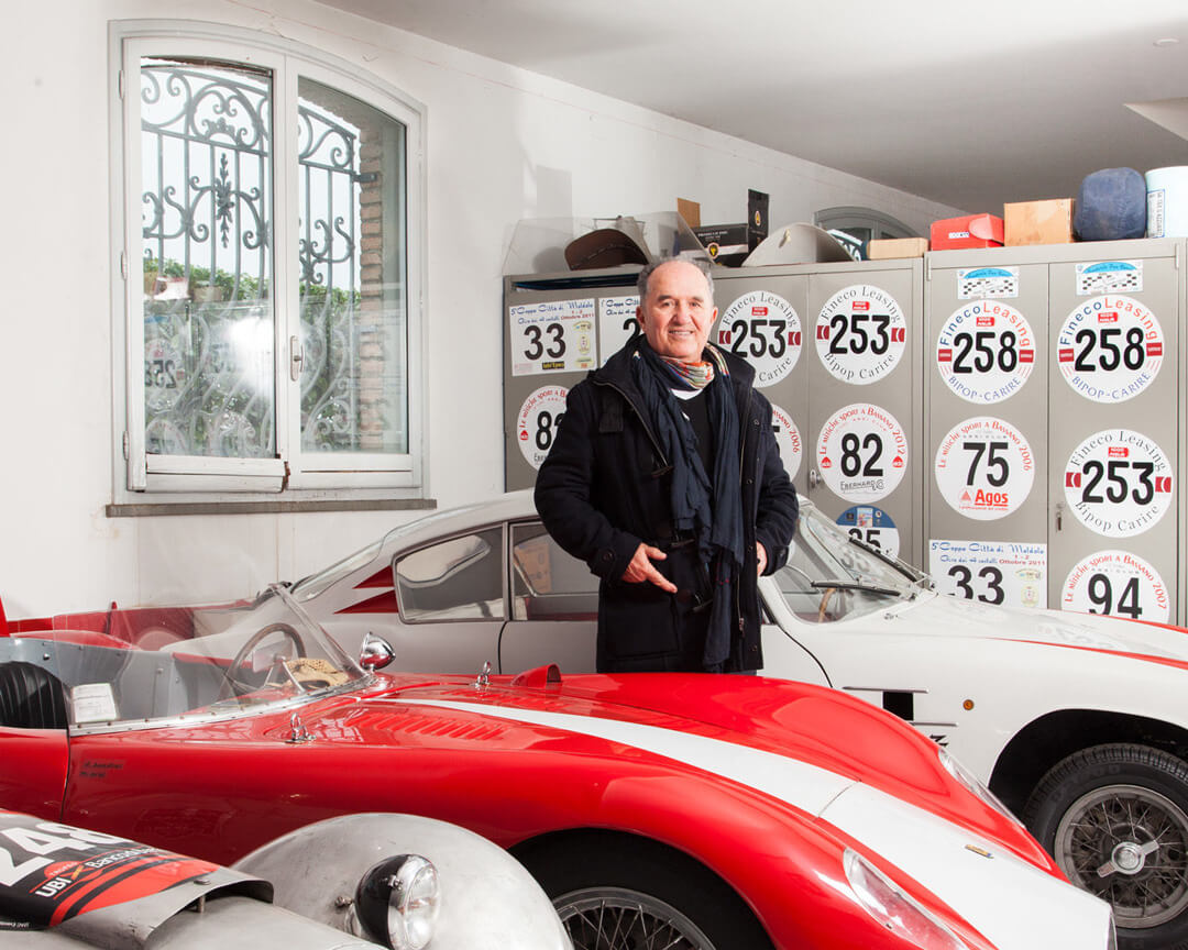 Foto di Ilario Bandini tra le sue auto d'epoca con alle spalle numeri appartenenti alle macchine
