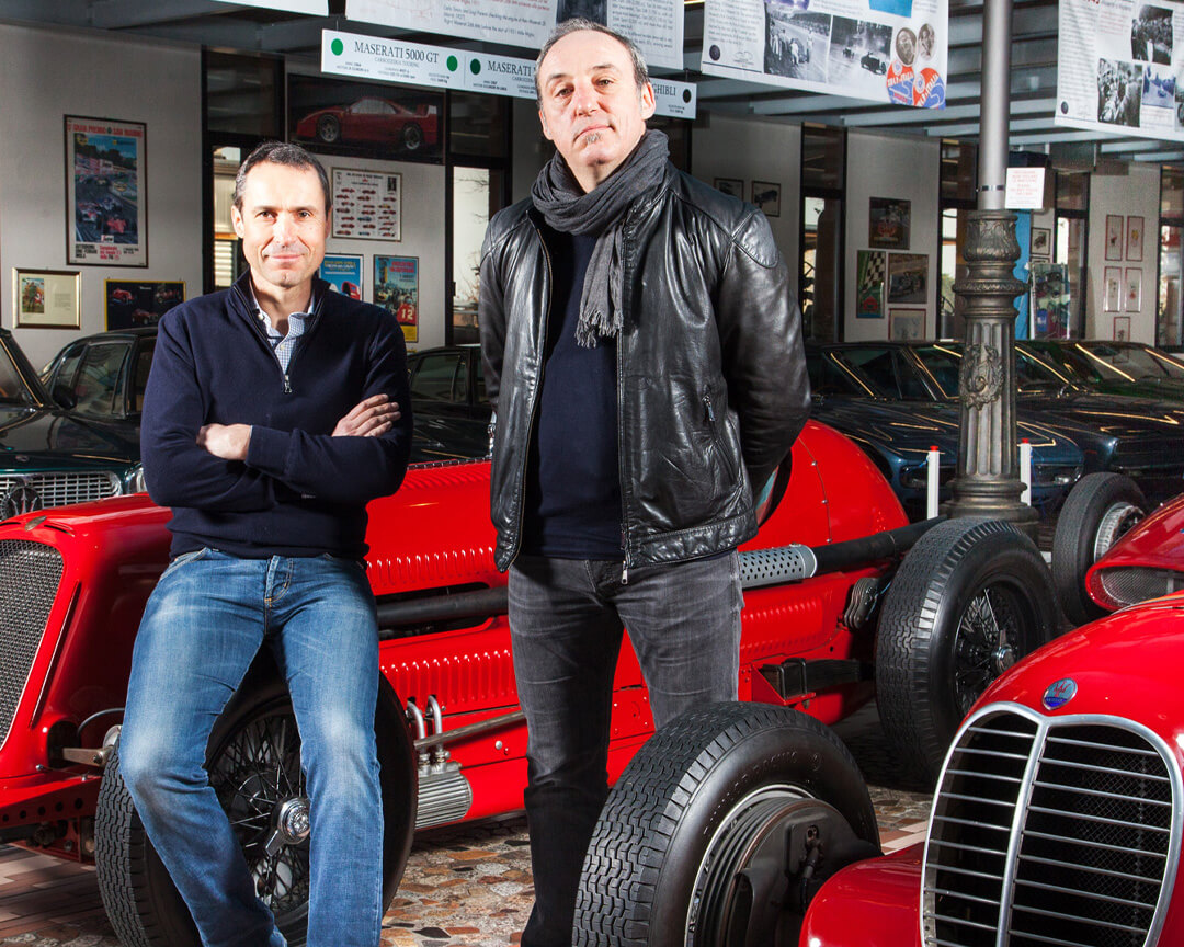 foto di Giovanni e Umberto Panini sorridenti con alle loro spalle la collezione di auto d'epoca