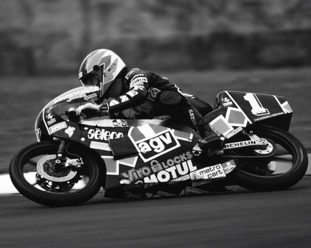 black-and-white photo of motogp rider leaning over a race bike, with livery full of sponsor logos such as AGV and Motul, as he takes a corner at high speed.