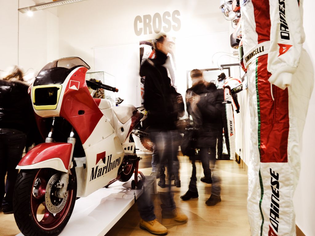 Interno di una sala del museo con visitatori in movimento; in primo piano a sinistra è esposta una moto da corsa bianca e rossa con sponsor Marlboro, mentre sulla destra si vede una tuta da pilota.