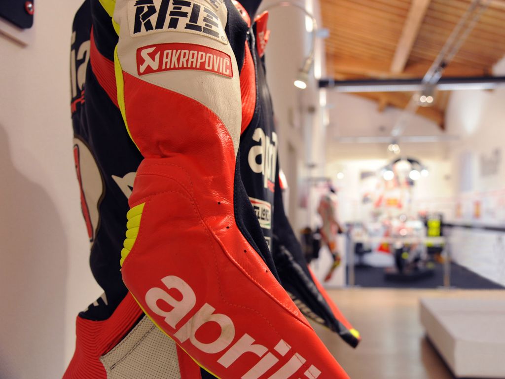 Dettaglio ravvicinato della manica di una tuta da motociclista in pelle rossa, nera e bianca, con i loghi degli sponsor Aprilia e Akrapovic ben visibili.