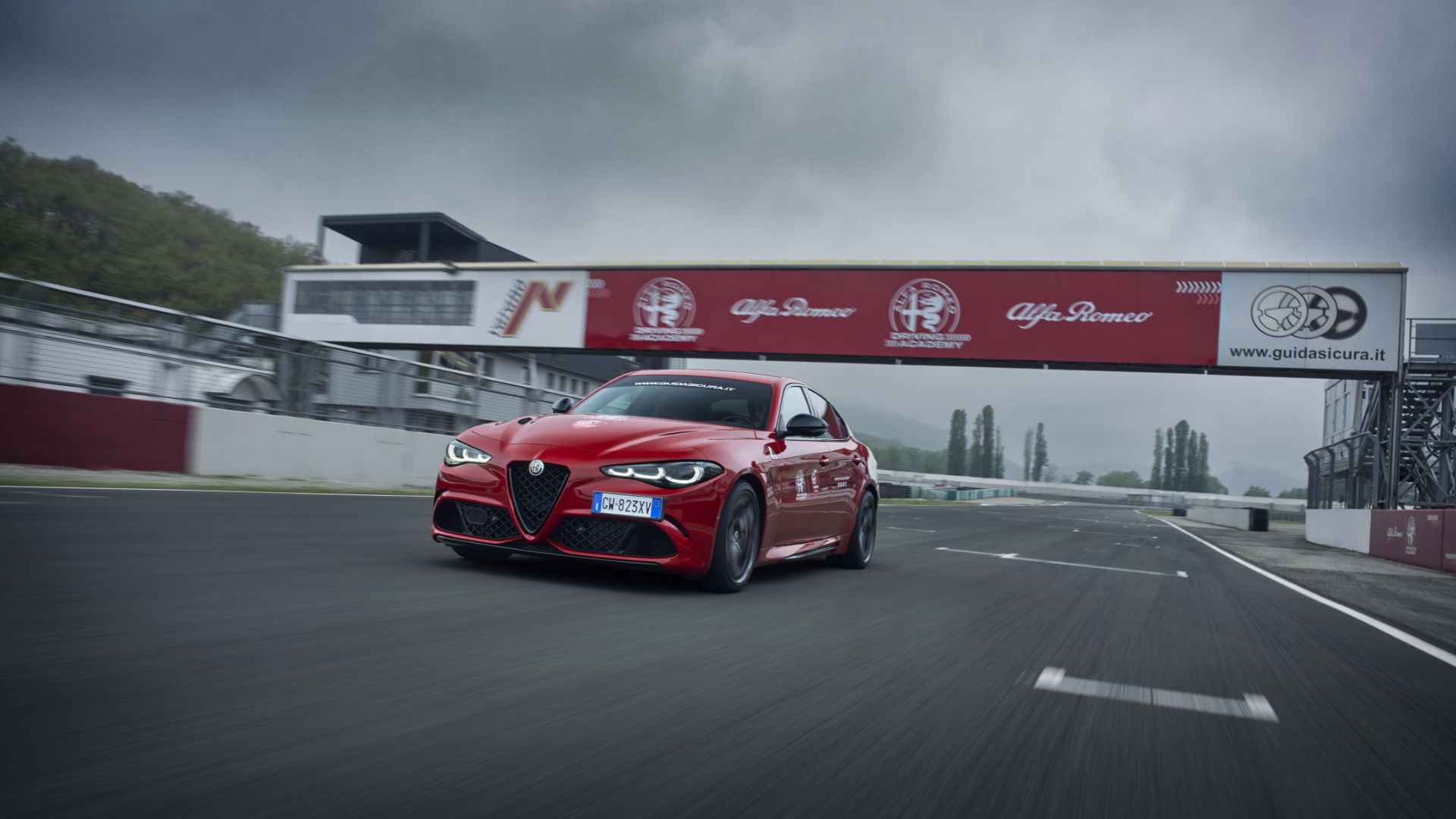 Un'Alfa Romeo Giulia rossa in accelerazione sul rettilineo di un circuito automobilistico, mentre passa sotto la struttura dell'Alfa Romeo Driving Academy sotto un cielo nuvoloso.