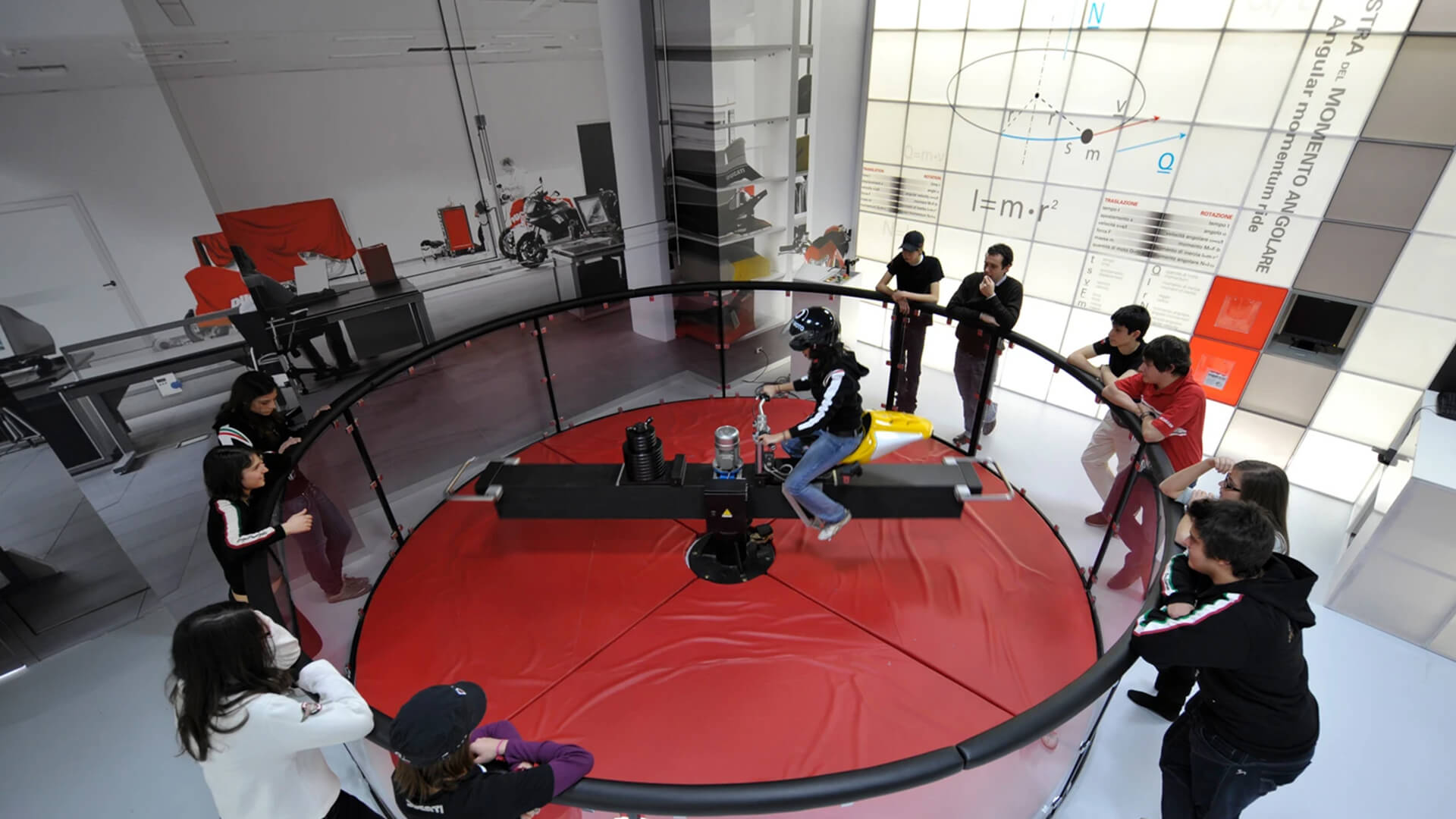 The image shows a group of people watching a motorcycle simulator, where a person wearing a helmet is sitting on a motorcycle mounted on a rotating platform