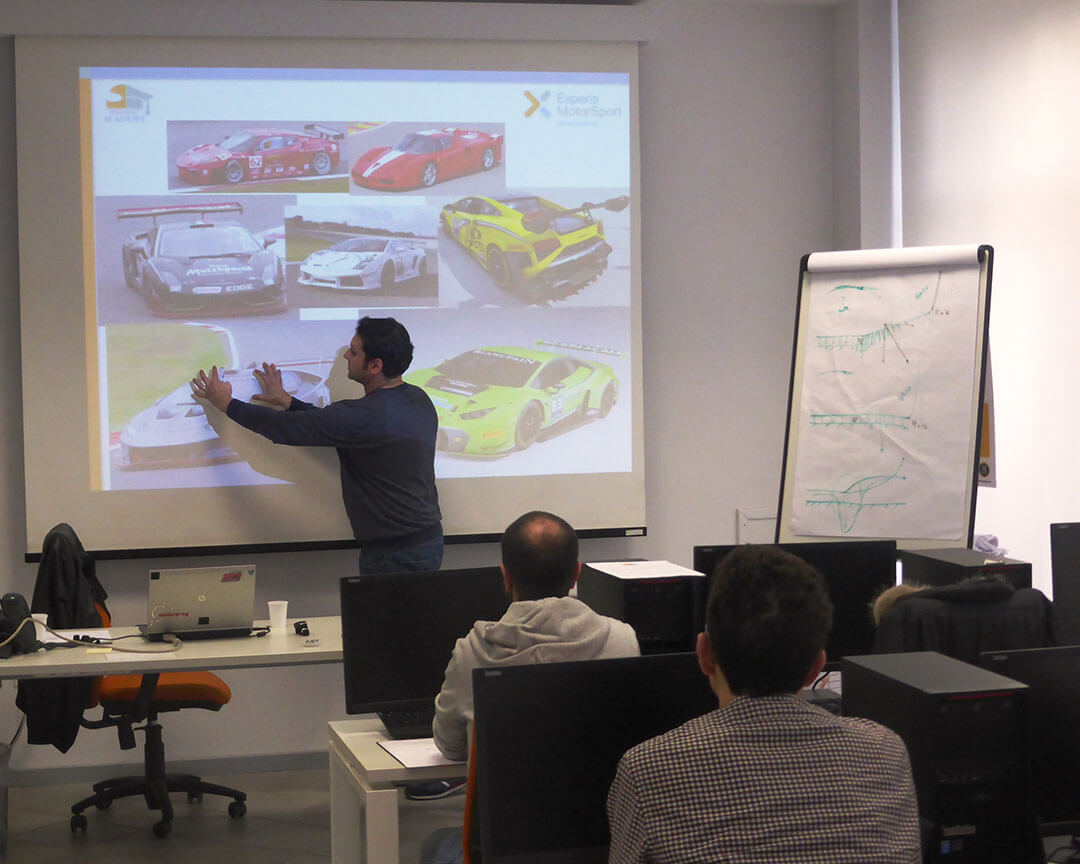 teacher pointing to the board on which images of racing cars are projected in front of 2 students sitting at their desks