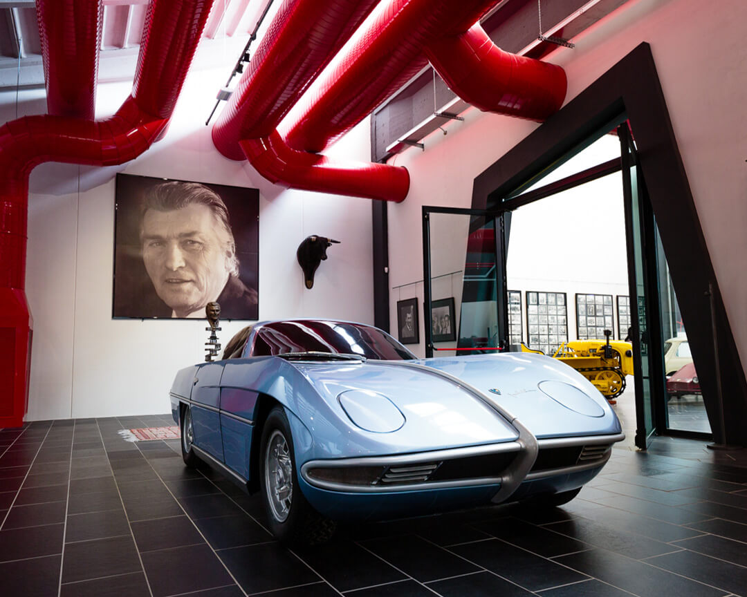 front of a vintage blue lamborghini car with portrait of ferruccio lamborghini behind it