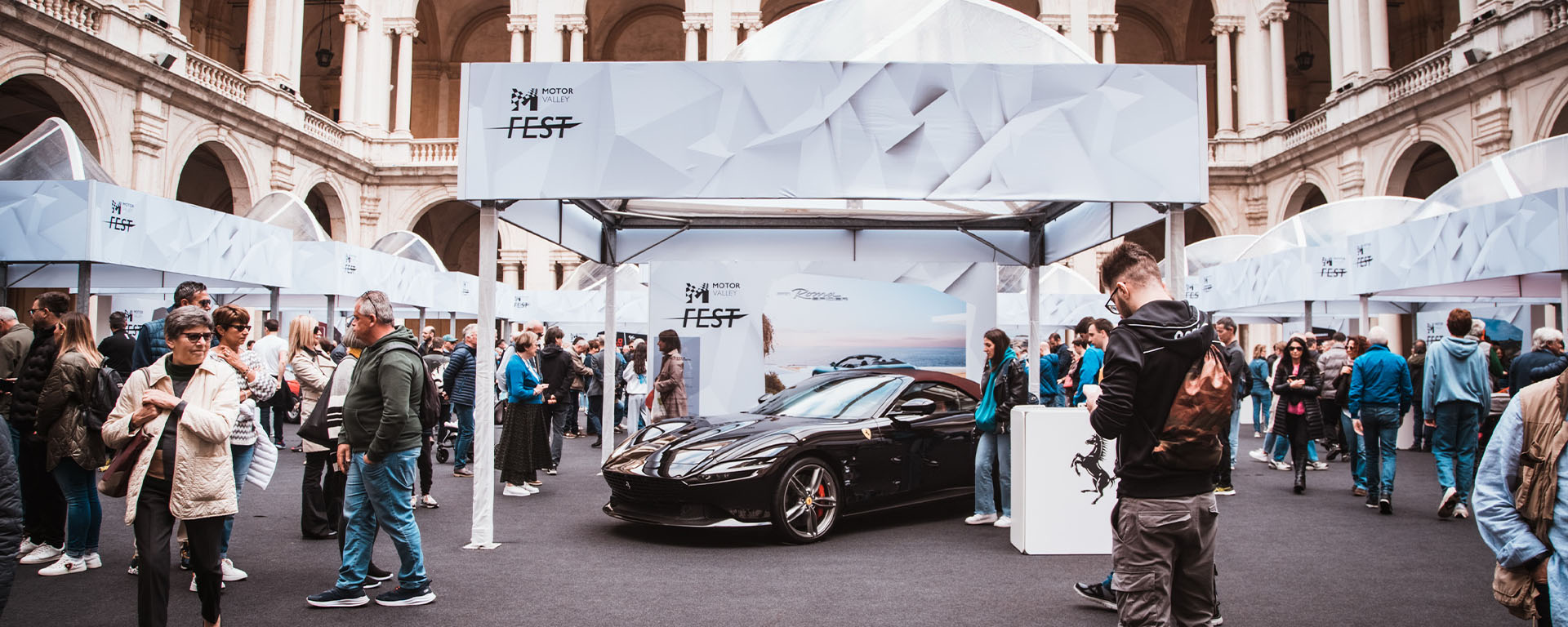 ferrari  nera esposta al Motor Valley fest sotto un gazebo con logo tra le persone che affollano il chiostro di un palazzo medioevale a Modena