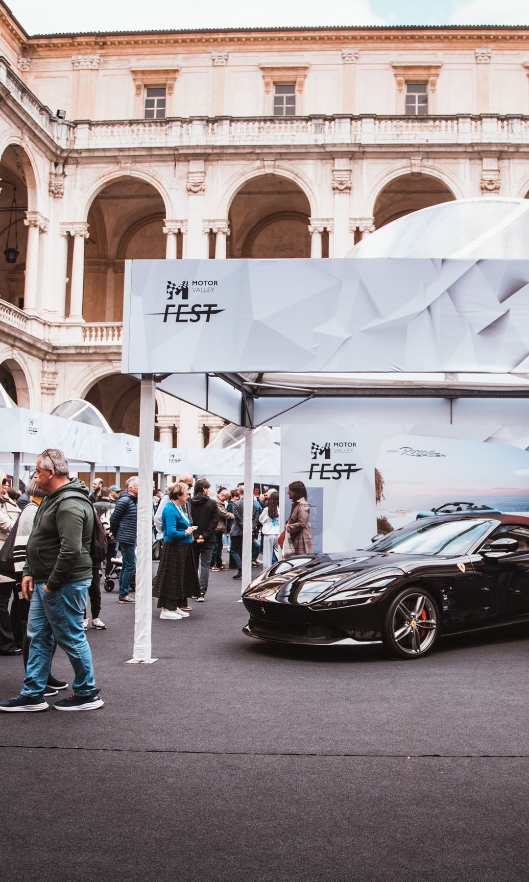 ferrari  nera esposta al Motor Valley fest sotto un gazebo con logo tra le persone che affollano il chiostro di un palazzo medioevale a Modena