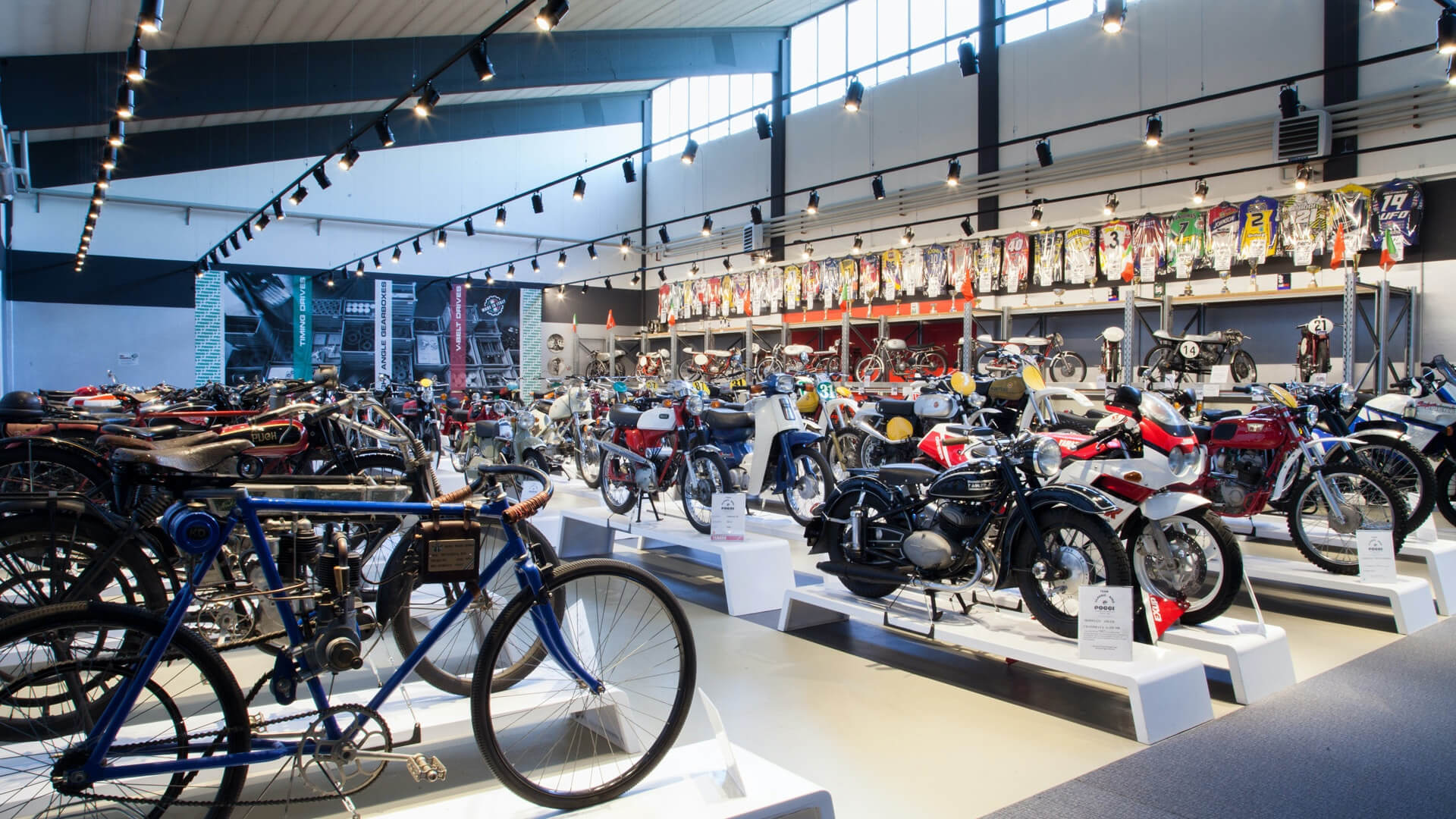 panoramica della sala con diverse moto e biciclette esposte e appese diverse tute di moto da collezione