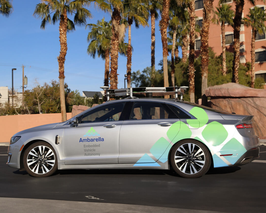 side view of grey car with Ambarella logo and coloured geometric shapes on the rear of the bodywork driving along a paved road with palm trees behind it