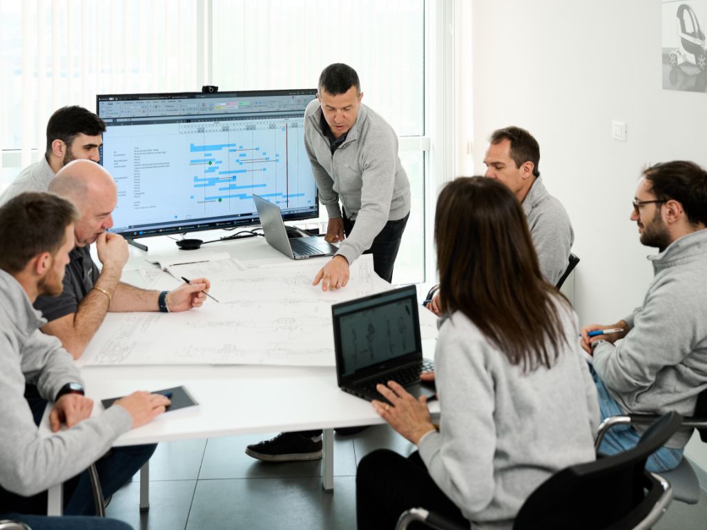 Scrivania di un ufficio con monitor, dipendenti seduti intorno ad una scrivania scrivono su un unico grande foglio, alcuni hanno il portatile.