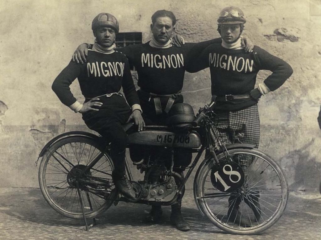 Tre piloti abbracciati in posa per una foto davanti a una moto d'epoca.