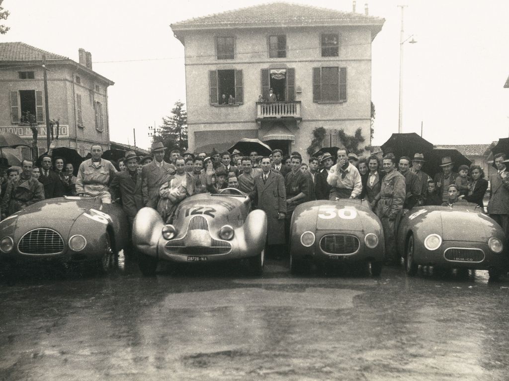 I piloti della Squadra Stanguellini con le rispettive automobili da corsa, in posa per una foto con i fan alle spalle, in una giornata di pioggia in piazza a Modena.