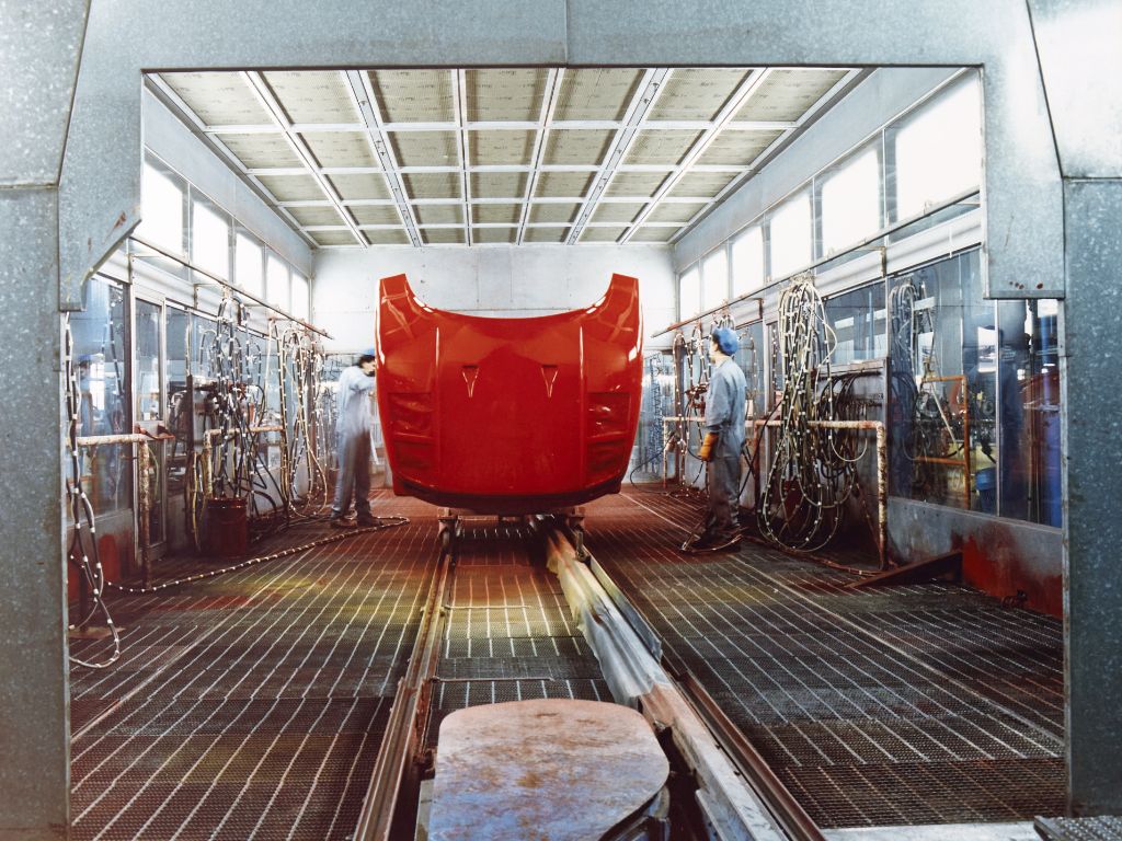 Cofano rosso di una Ferrari in fase di verniciatura, con due operai al lavoro su di esso, all'intero di una fabbrica.