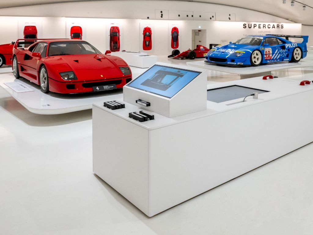 Schermo interattivo di un museo e sullo sfondo Ferrari esposte su piedistalli.