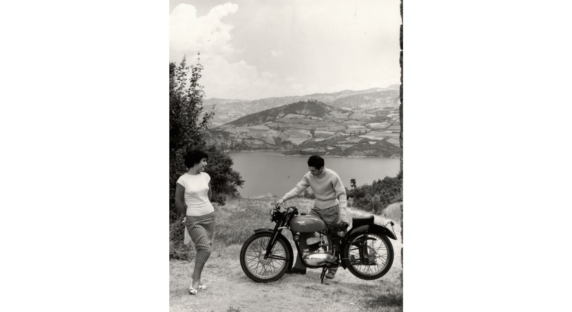 Foto d'epoca di una giovane donna a fianco di un giovane uomo con la sua motocicletta. Sullo sfondo un paesaggio collinare ed un lago.