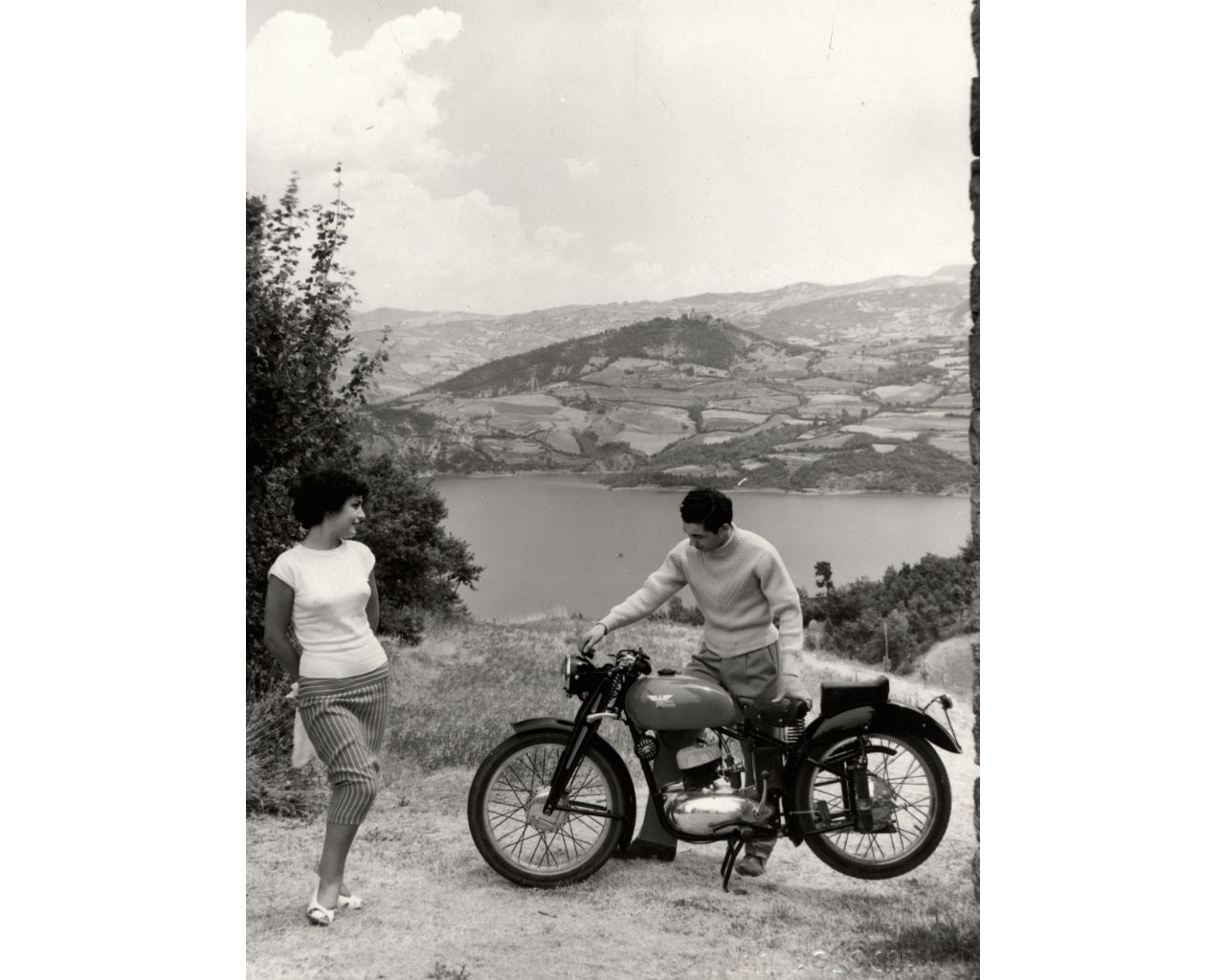 Foto d'epoca di una giovane donna a fianco di un giovane uomo con la sua motocicletta. Sullo sfondo un paesaggio collinare ed un lago.