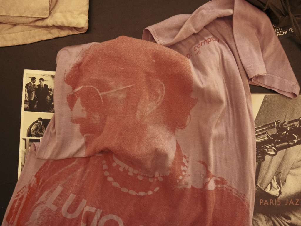 Maglietta rosa di C.P. Company con una stampa di Lucio Dalla, sopra un tavolo con alcune fotografie e riviste.