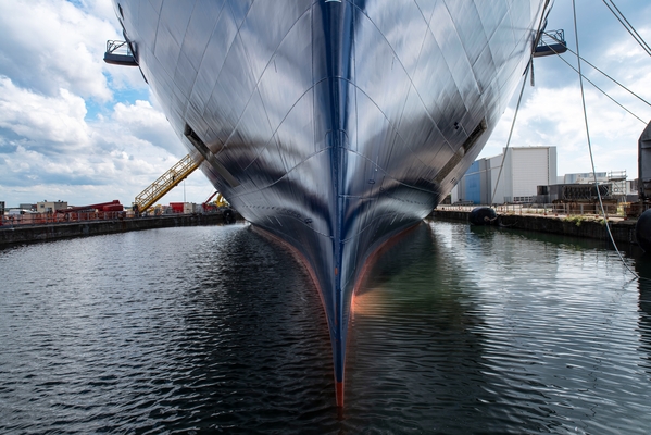 Vista prospettica della prua imponente di una grande nave blu in un bacino di carenaggio, con lo scafo che taglia la superficie dell'acqua.