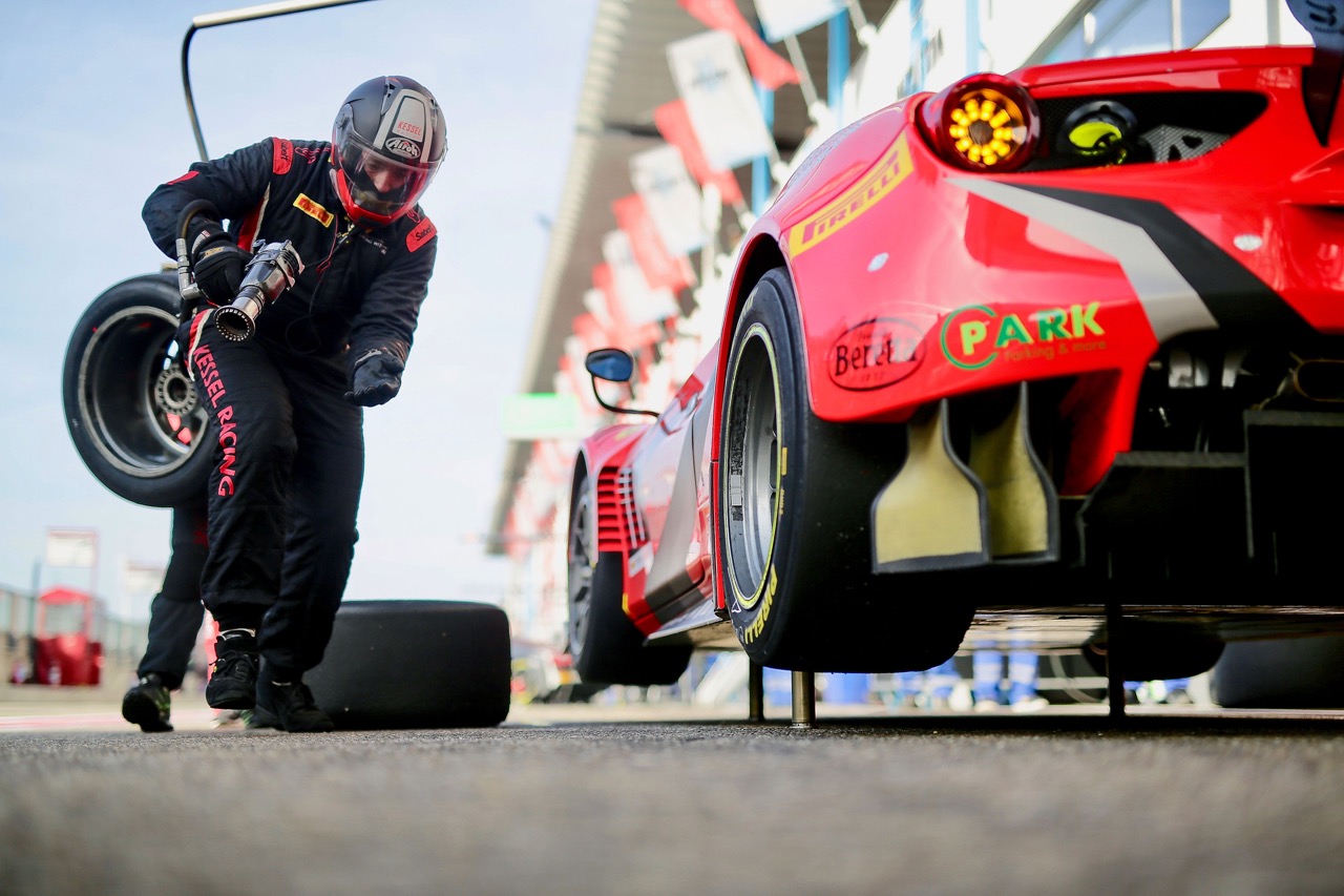 Un meccanico in tuta e casco solleva uno pneumatico da corsa accanto a una Ferrari 488 GT3 sollevata sui martinetti.
