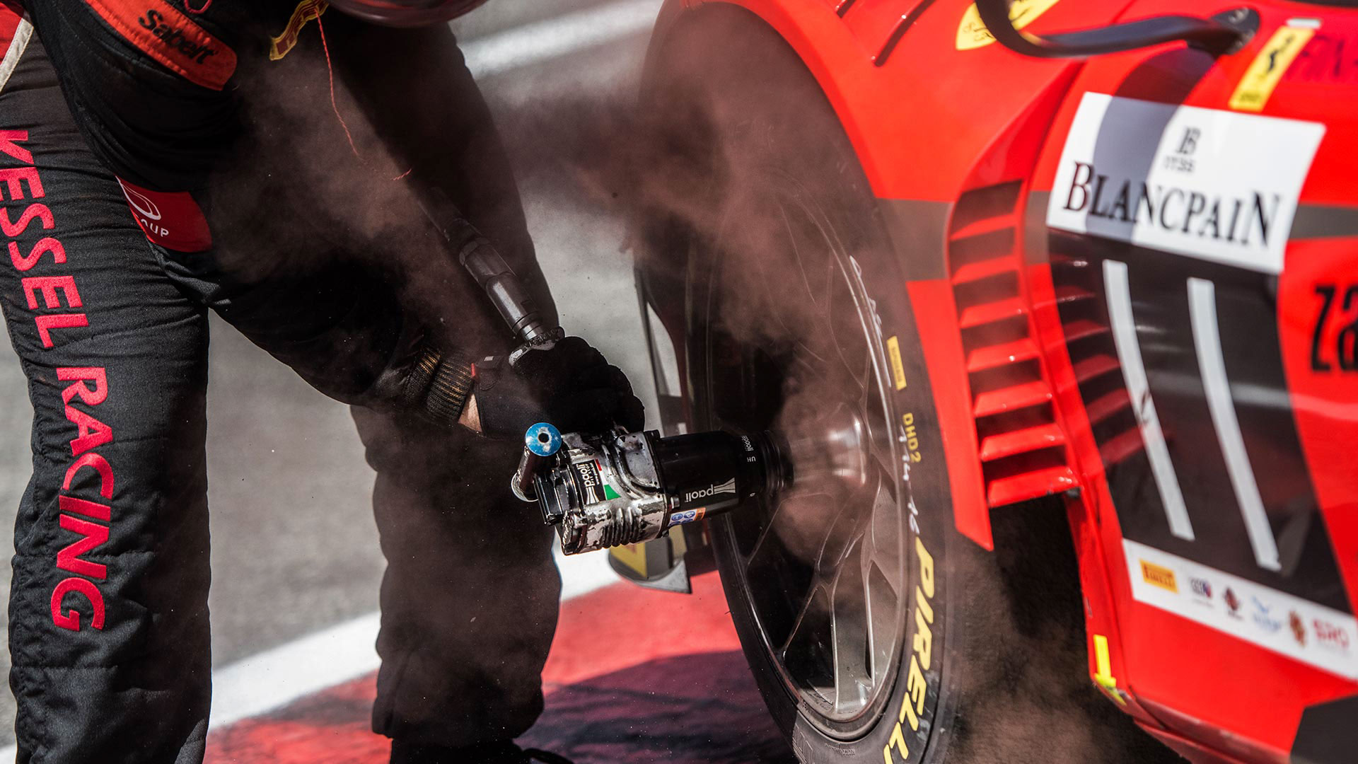 Dettaglio di un avvitatore a impulsi in azione sulla ruota di un'auto da corsa rossa, con polvere di frenata che si solleva per la velocità.