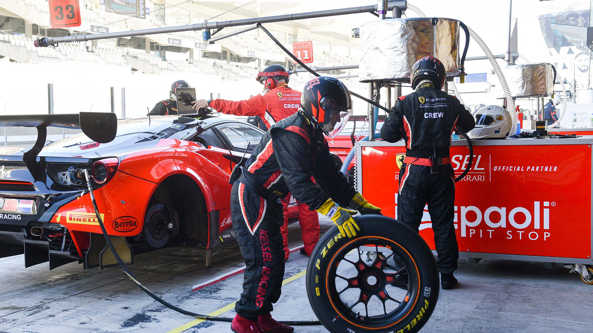 Scena frenetica in corsia box con meccanici che sostituiscono gli pneumatici di una Ferrari rossa presso una postazione Paoli Pit Stop.
