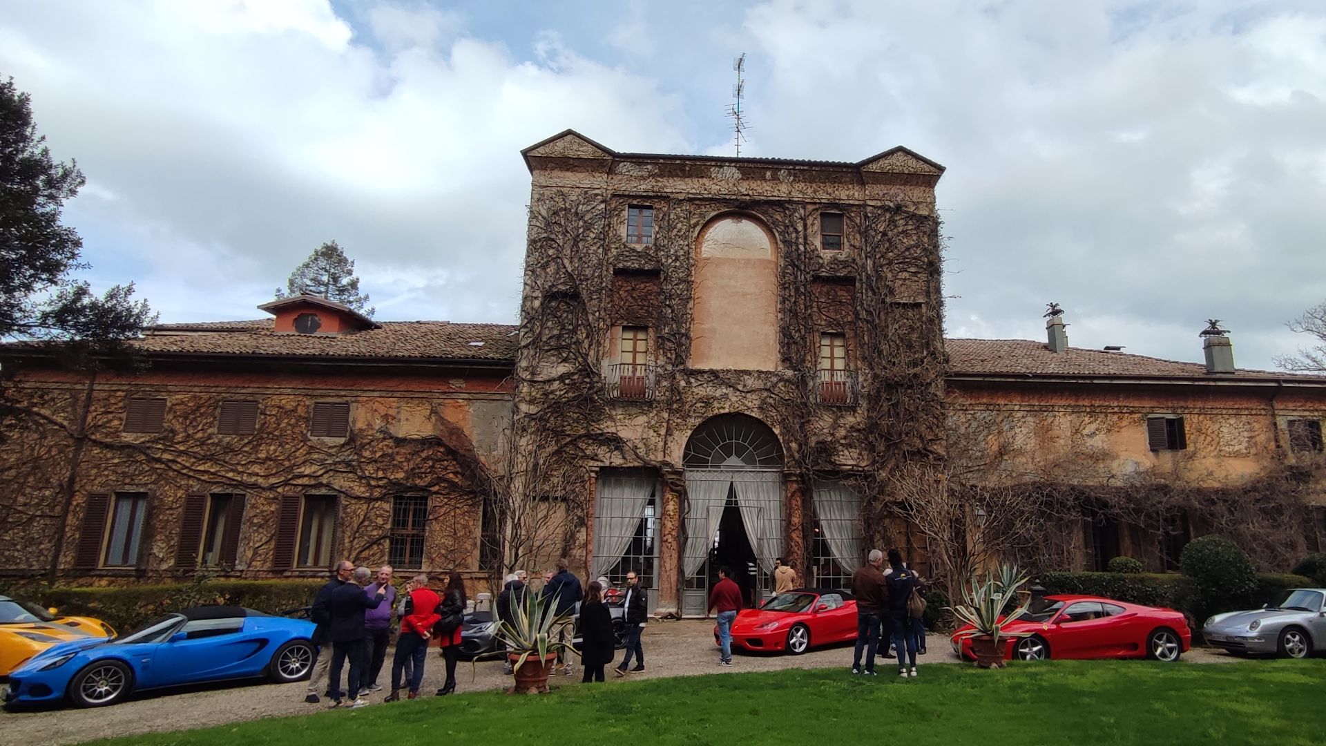 Auto storiche di lusso parcheggiate di fronte a una villa storica emiliana, con della gente intorno.