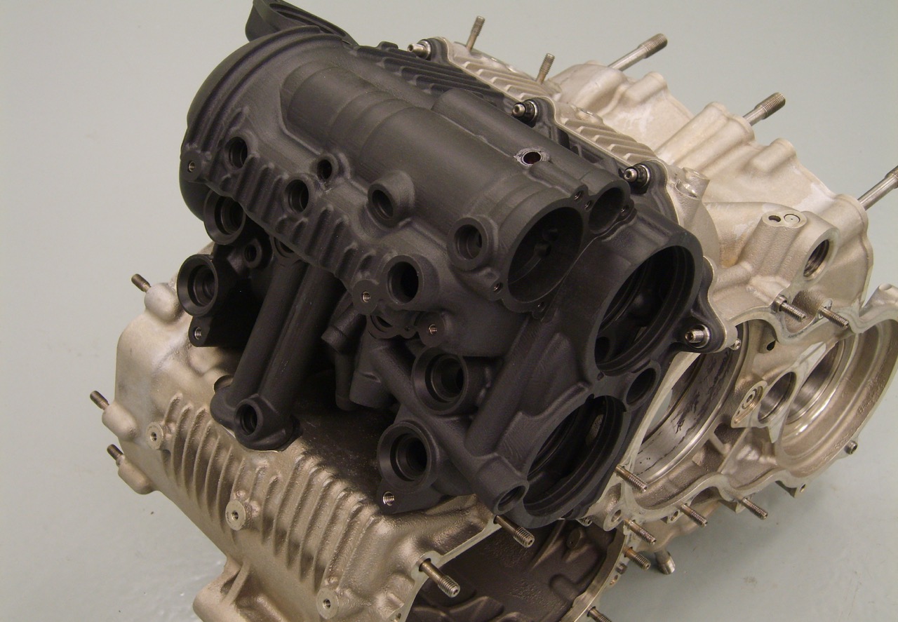 Componente meccanico complesso stampato in 3D in materiale nero opaco, montato su una base metallica di un motore.