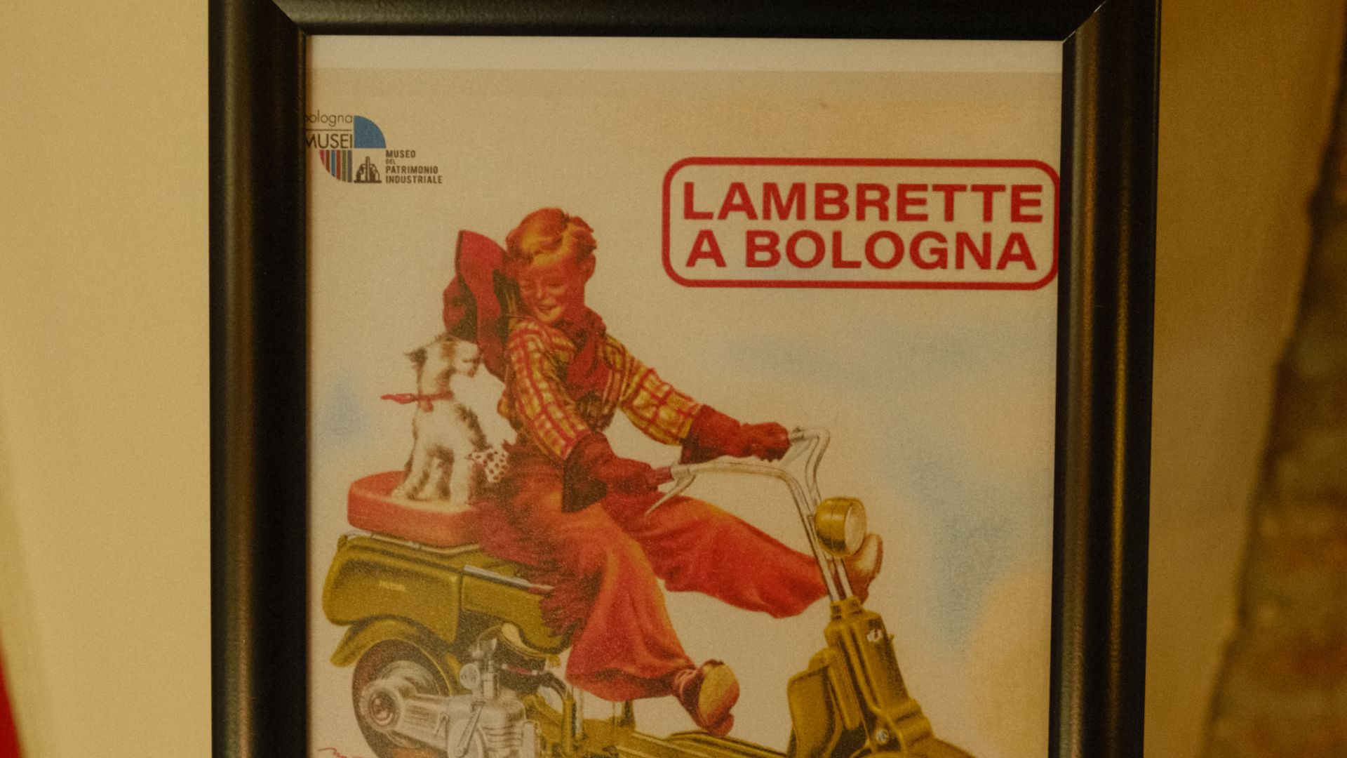 Illustrazione vintage incorniciata di un ragazzo e un cane su uno scooter con la scritta "Lambrette a Bologna", presso il Museo del Patrimonio Industriale.