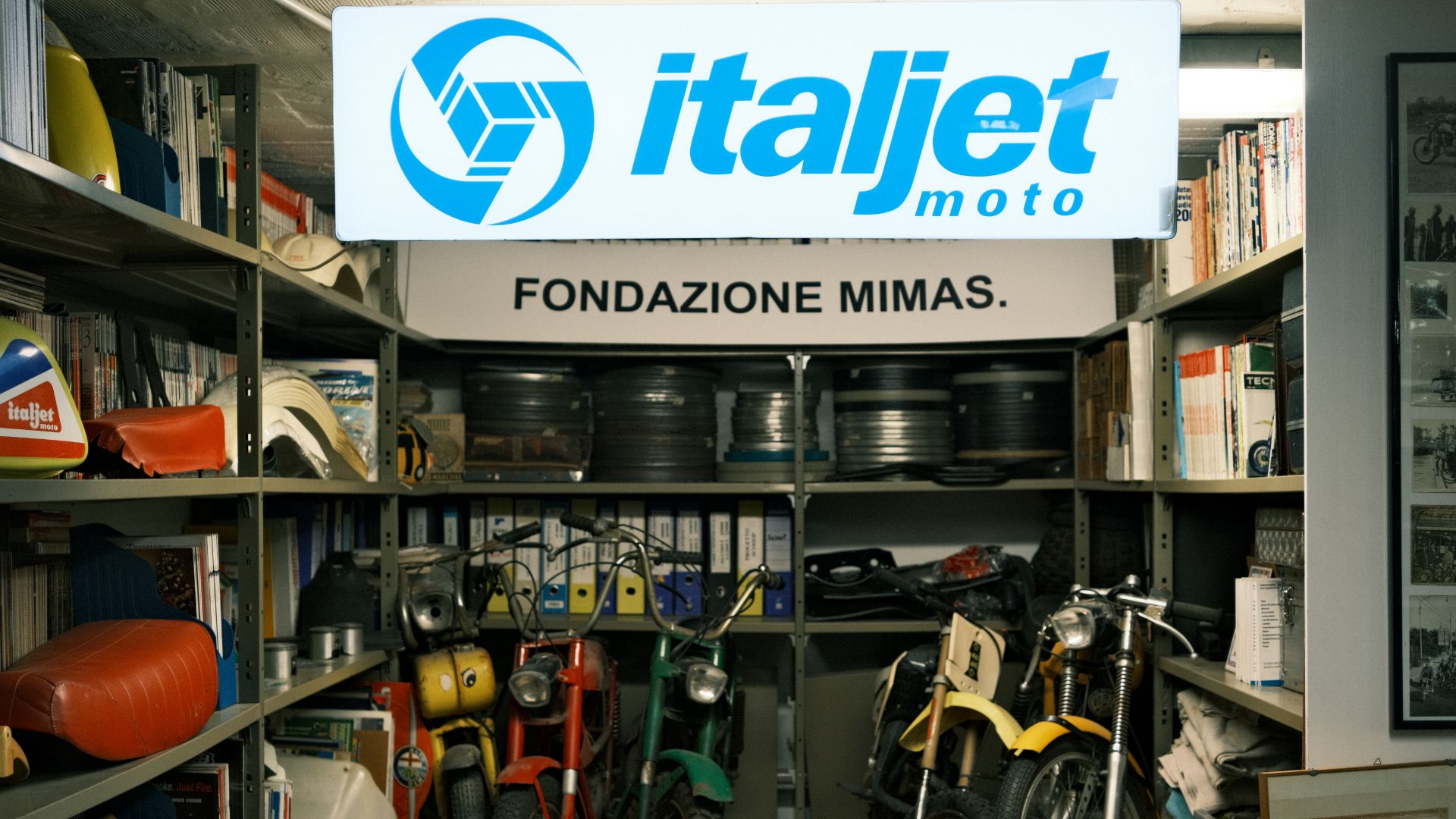 Magazzino con scaffali pieni di documenti, rulli di pellicola e motorini d'epoca Italjet.