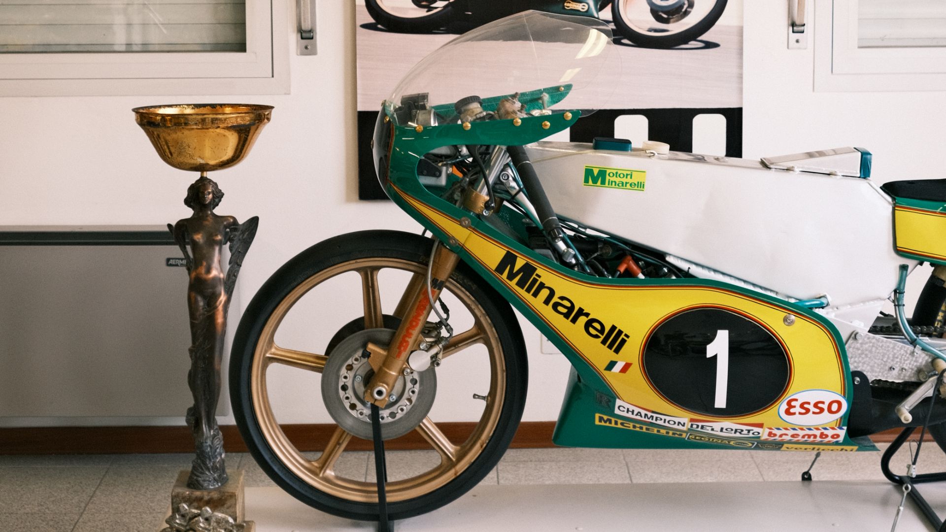 Primo piano di una moto da corsa Minarelli verde e gialla con il numero 1, accanto a un trofeo dorato.