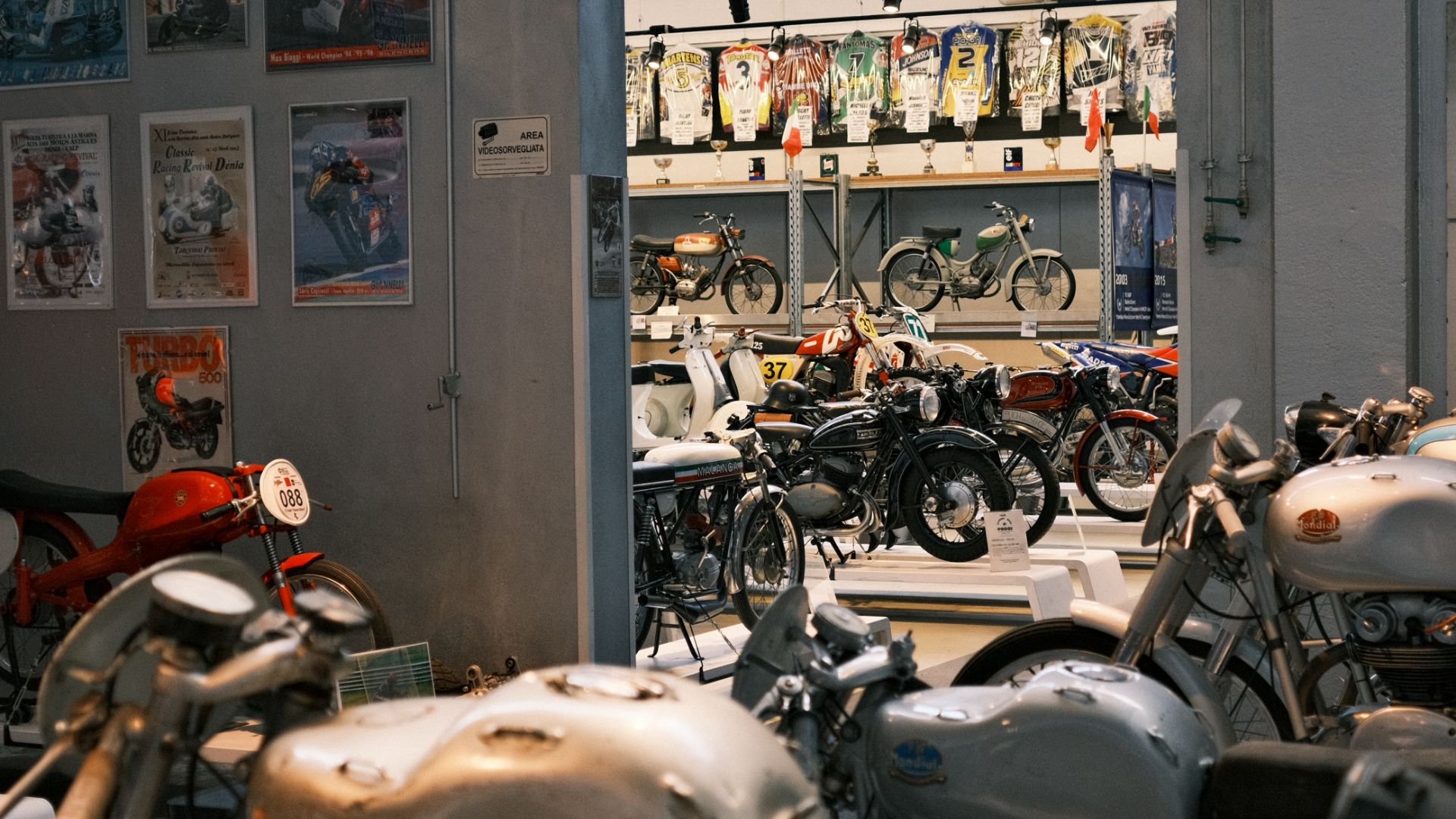 Interno della Collezione Moto Poggi con diverse file di motociclette storiche e poster vintage alle pareti.