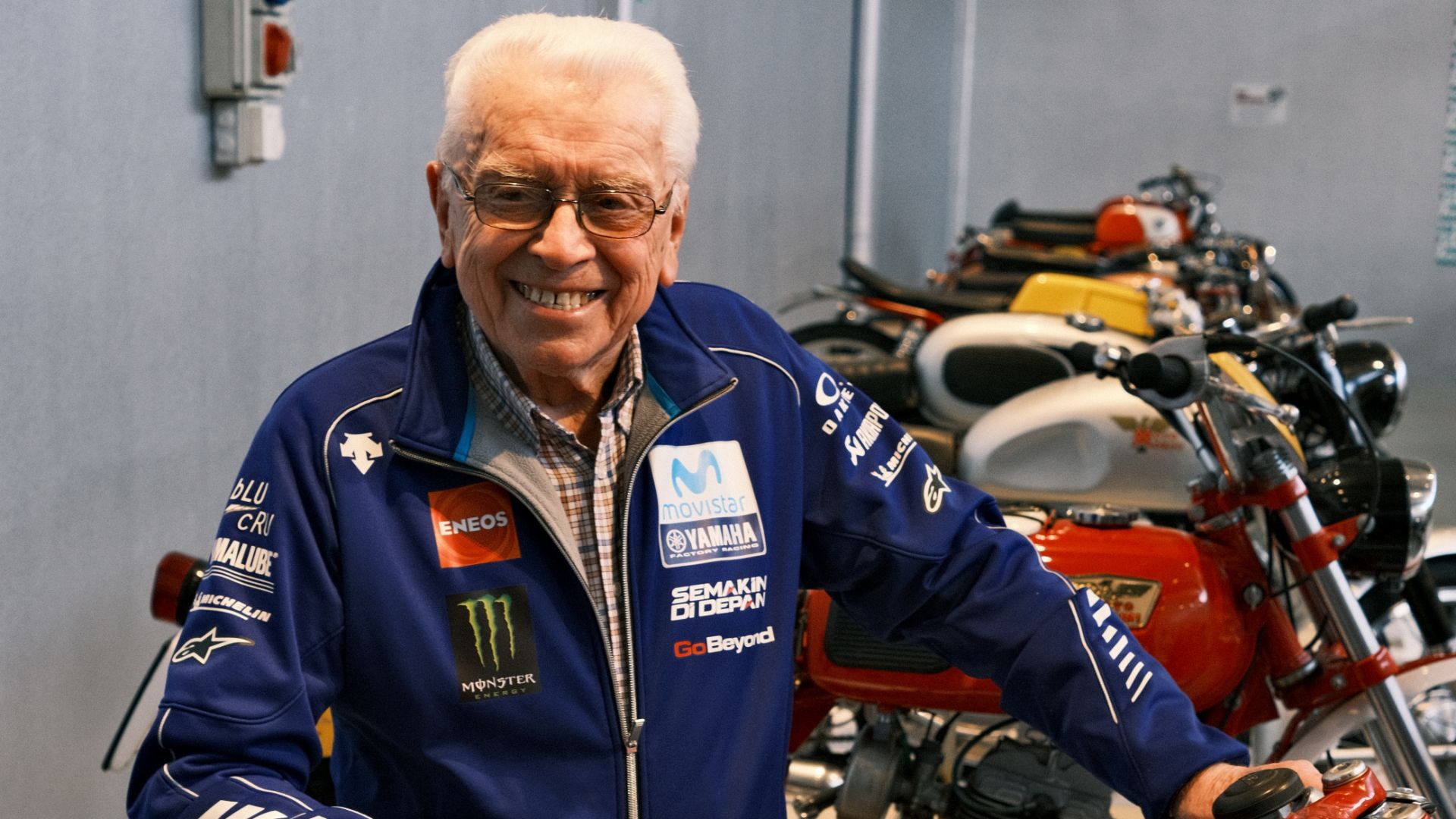 Un uomo anziano sorridente con occhiali e giacca blu Yamaha Racing davanti a una fila di moto presso la Collezione Poggi.