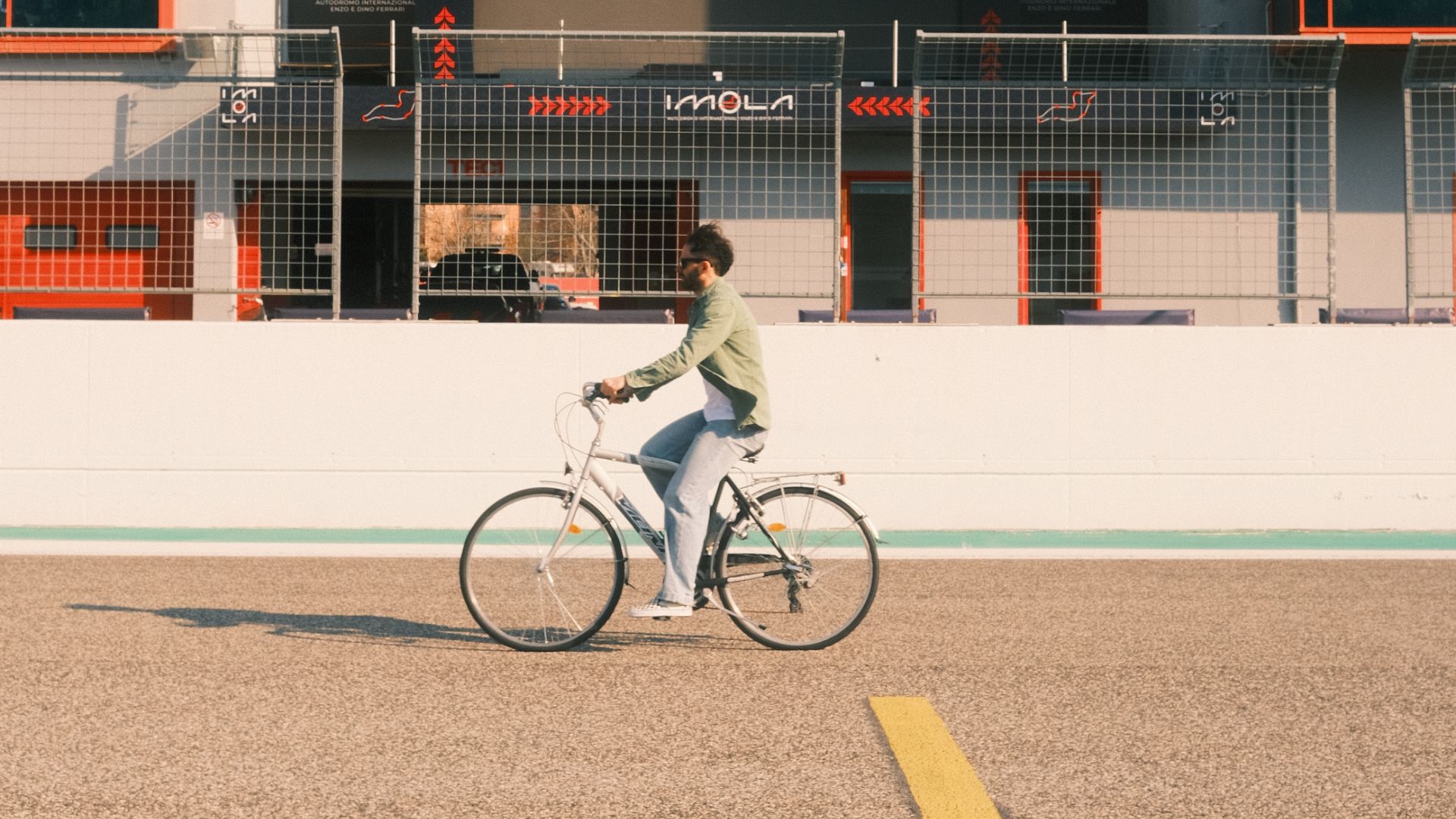 Un uomo attraversa in bicicletta il rettilineo dell'Autodromo Enzo e Dino Ferrari di Imola, con i box e i loghi del circuito sullo sfondo.