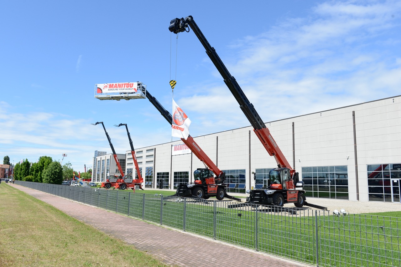 Esposizione esterna di sollevatori telescopici Manitou con diversi accessori montati: verricello con bandiera aziendale, piattaforma porta-persone e forche per pallet.
