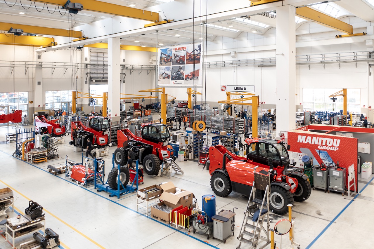 Veduta dell'ampia e moderna linea di assemblaggio all'interno dello stabilimento Manitou, con sollevatori telescopici in fase di montaggio e carriponte industriali.