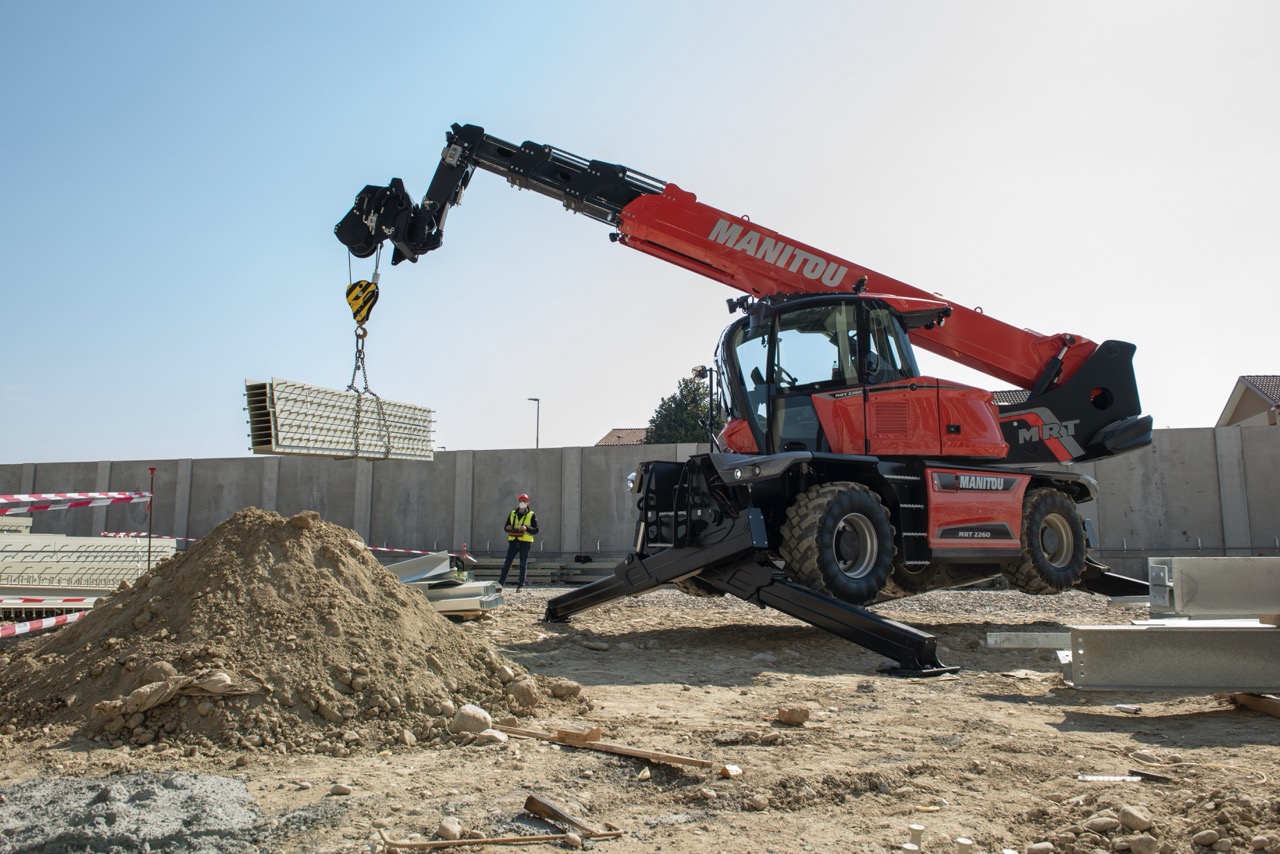 Sollevatore telescopico rotativo Manitou MRT 2260 che solleva un carico mediante gancio in un cantiere edile, con stabilizzatori estesi per la massima sicurezza.