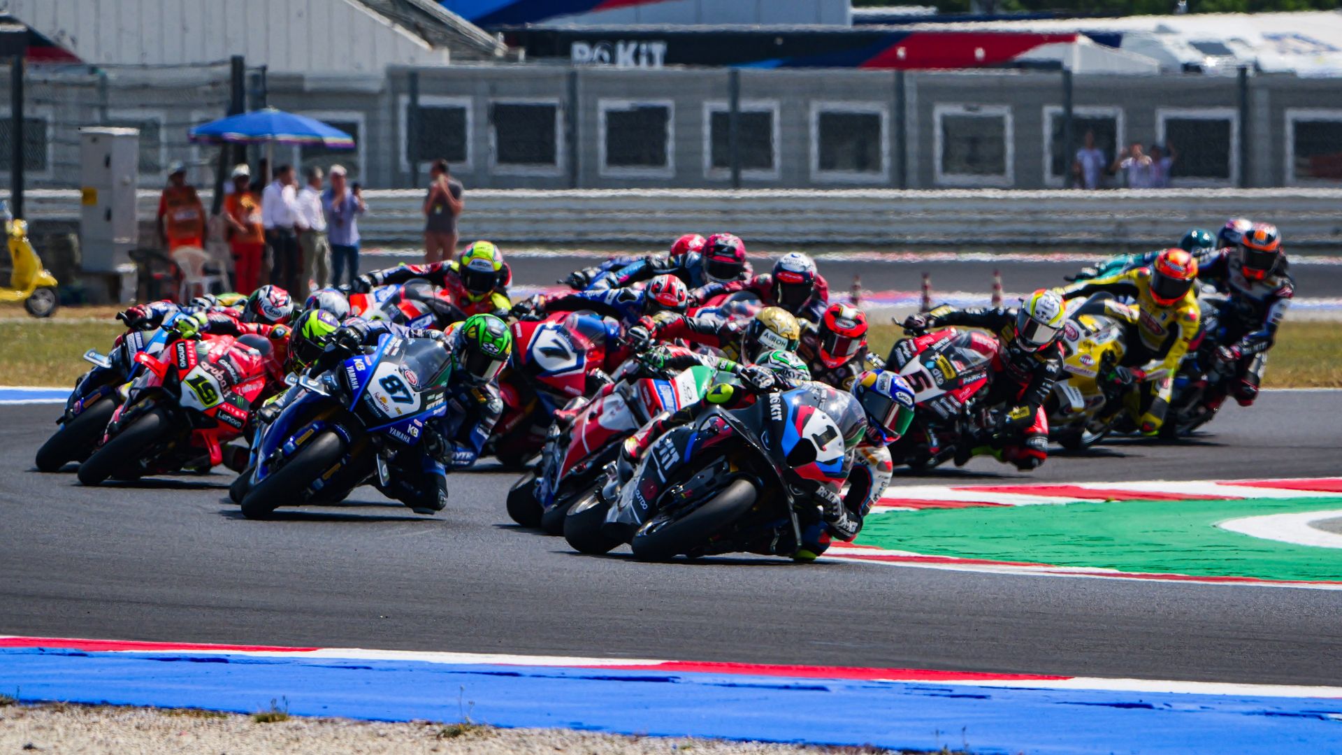 Un folto gruppo di piloti professionisti di Superbike in forte piega mentre affronta una curva a destra su un circuito, mostrando un momento di gara molto serrato e competitivo.