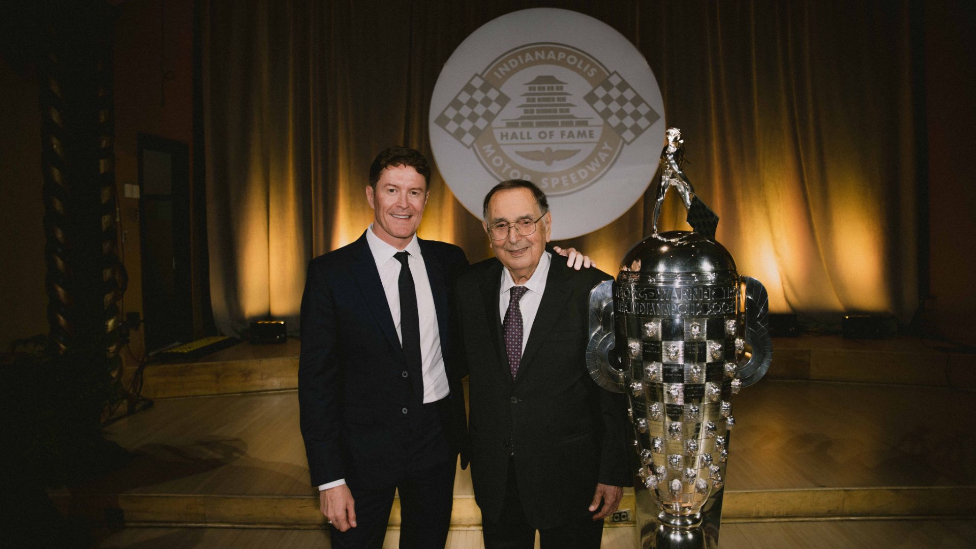 Scott Dixon e Giampaolo Dallara sorridono in posa su un palco accanto al monumentale Trofeo Borg-Warner d'argento, con lo sfondo del logo della Indianapolis Motor Speedway Hall of Fame.