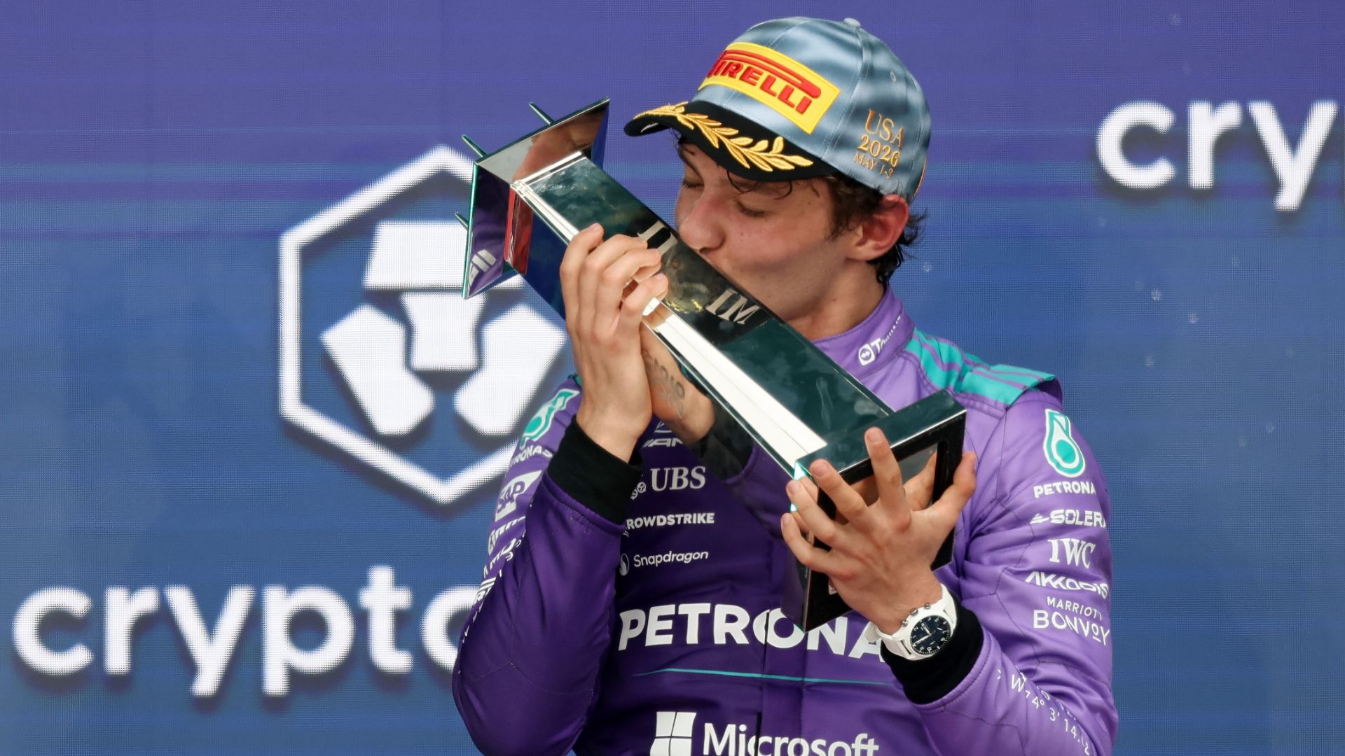Il pilota Kimi Antonelli, con indosso la tuta viola del team Mercedes e un cappellino celebrativo Pirelli del GP di Miami 2026, bacia il trofeo del vincitore tenendolo con entrambe le mani. Sullo sfondo si vede il logo Crypto.com.
