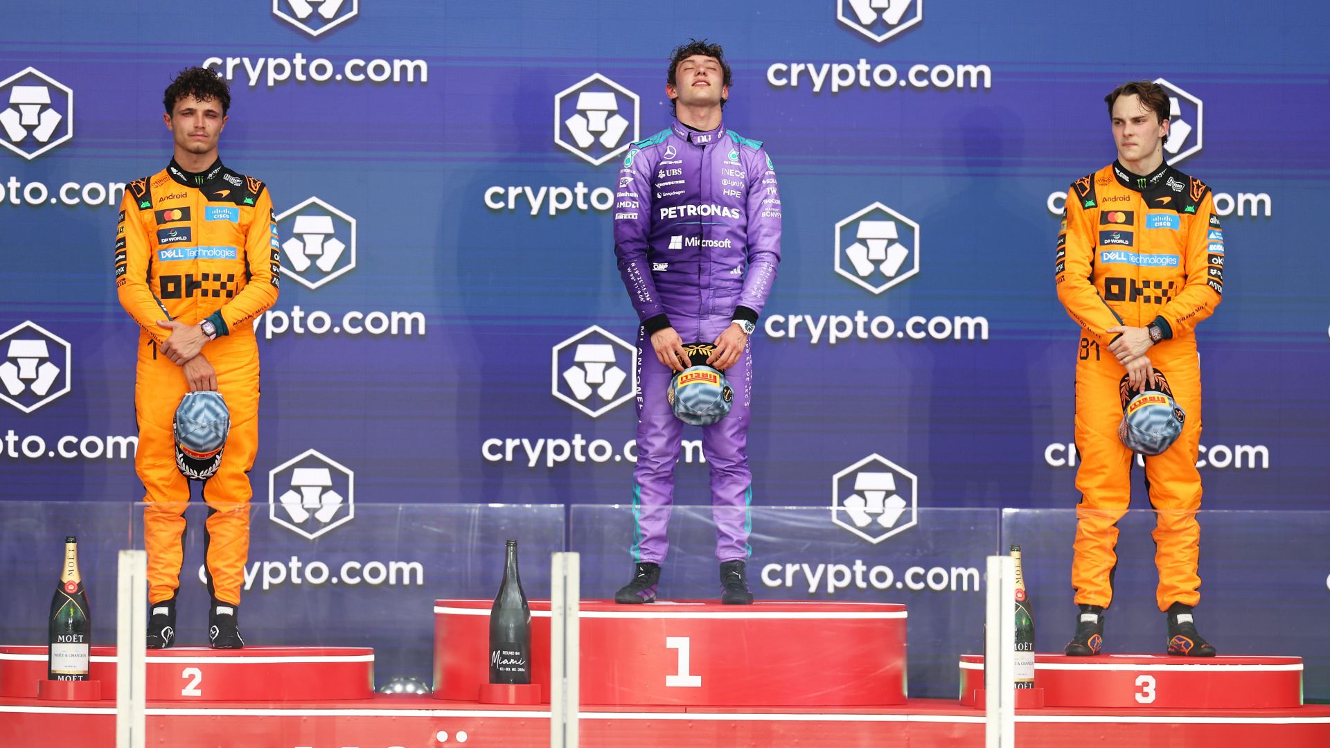 Podio del Gran Premio di Miami. Al centro, sul gradino più alto, Kimi Antonelli in tuta viola Mercedes con gli occhi chiusi in un momento di emozione. Ai lati, sui gradini 2 e 3, i piloti McLaren Lando Norris e Oscar Piastri in tuta arancione. Davanti a loro le bottiglie di champagne.