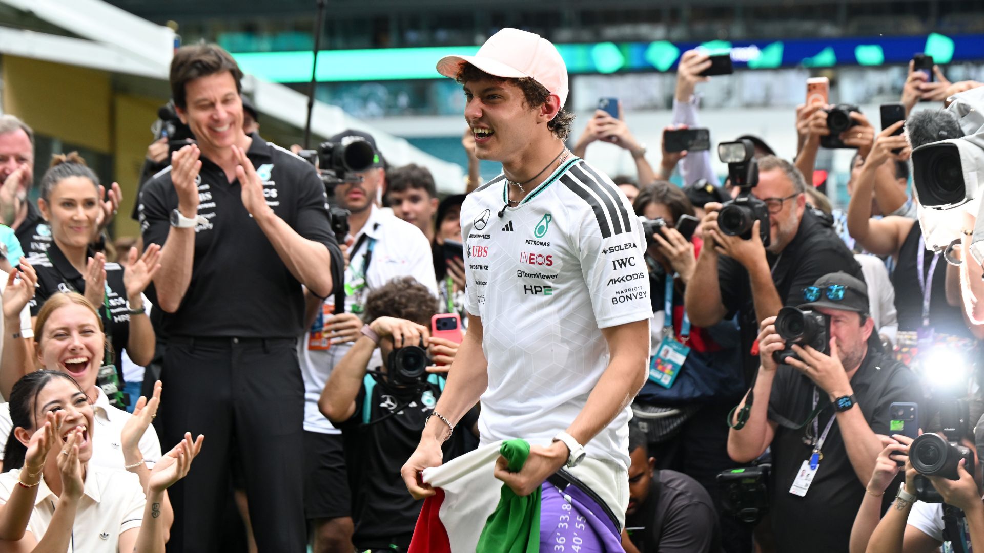 Kimi Antonelli cammina sorridente tra la folla e i fotografi nel paddock, portando una bandiera italiana sulle spalle. Indossa una maglia bianca Mercedes e un cappellino rosa. Alla sua sinistra, Toto Wolff sorride e applaude.
