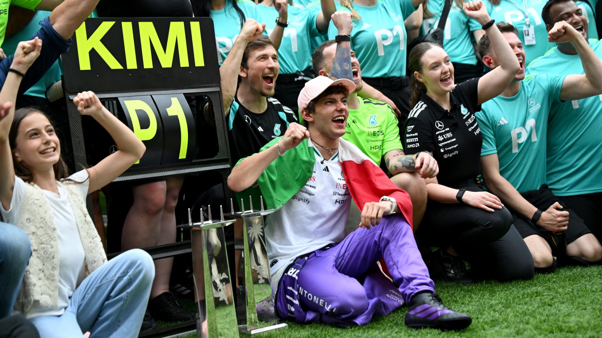 Kimi Antonelli seduto sul prato circondato dai membri del team Mercedes in maglia verde petrolio. Antonelli indossa la bandiera italiana come un mantello ed esulta con un pugno alzato. Accanto a lui, un tabellone con la scritta "KIMI P1".