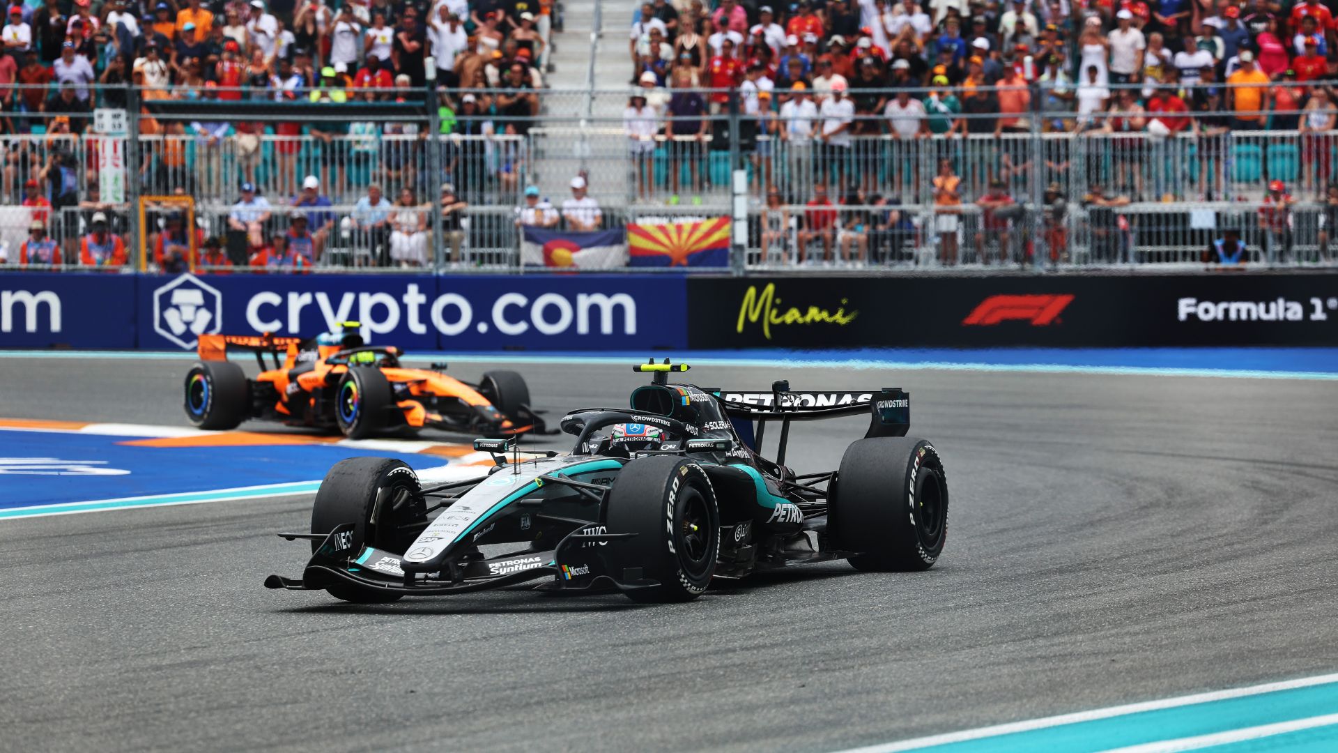La Mercedes nera e argento di Kimi Antonelli sfreccia su una curva del circuito di Miami. Inseguito a breve distanza da una McLaren arancione. Sullo sfondo si vedono le tribune affollate e i cartelloni pubblicitari di Crypto.com e F1.