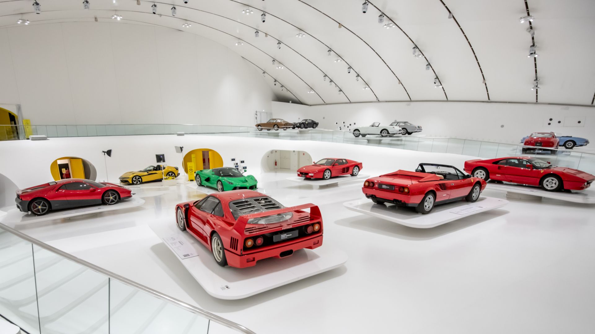 Panoramica dell'interno del Museo Enzo Ferrari di Modena con diverse supercar e auto storiche esposte nel grande padiglione bianco.