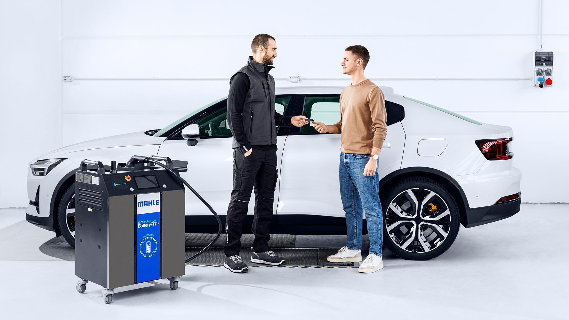 Un meccanico consegna le chiavi a un cliente all'interno dell'officina davanti a un'auto bianca, mentre in primo piano a sinistra spicca la stazione di ricarica e diagnostica MAHLE E-CHARGE 20 BatteryPRO.