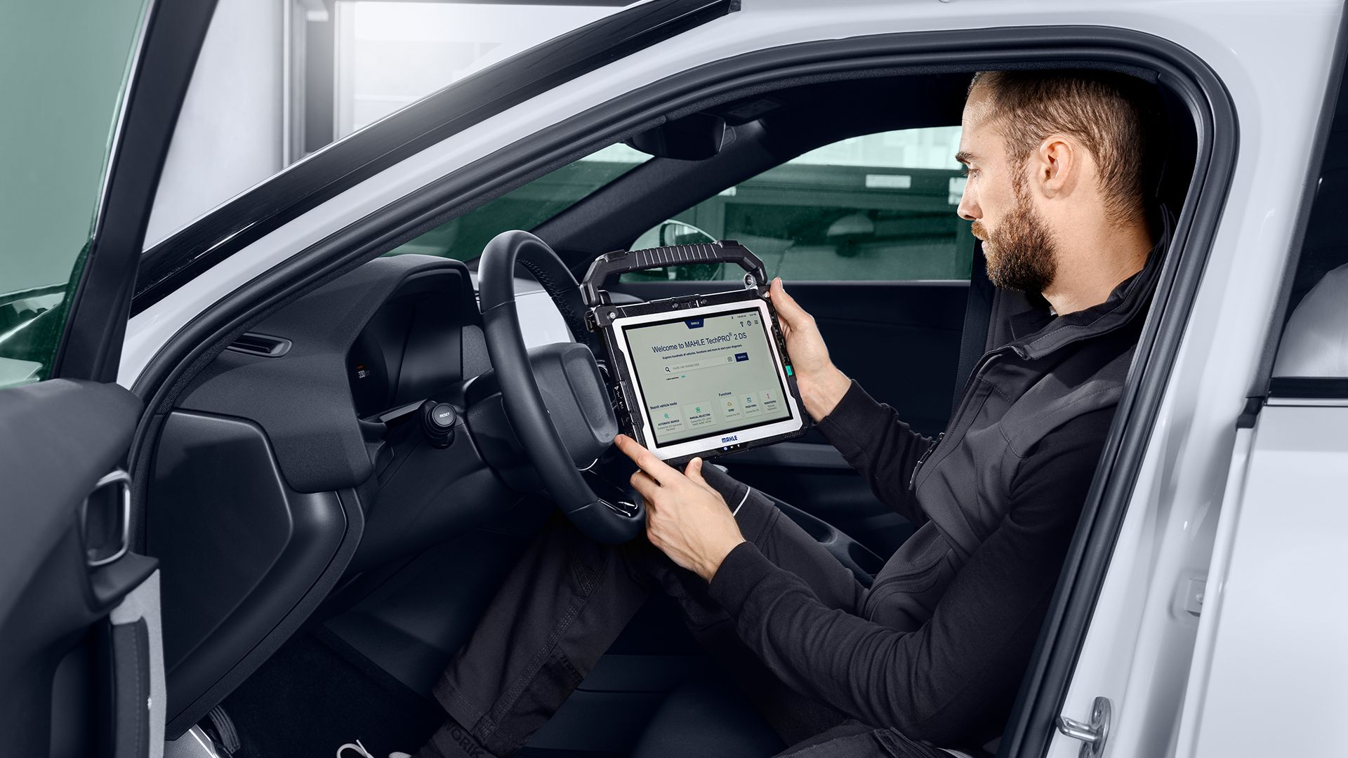 Un meccanico seduto all'interno del posto di guida di un'auto bianca mentre tiene in mano e consulta il tablet diagnostico MAHLE TechPRO 2 DS posizionato davanti al volante.