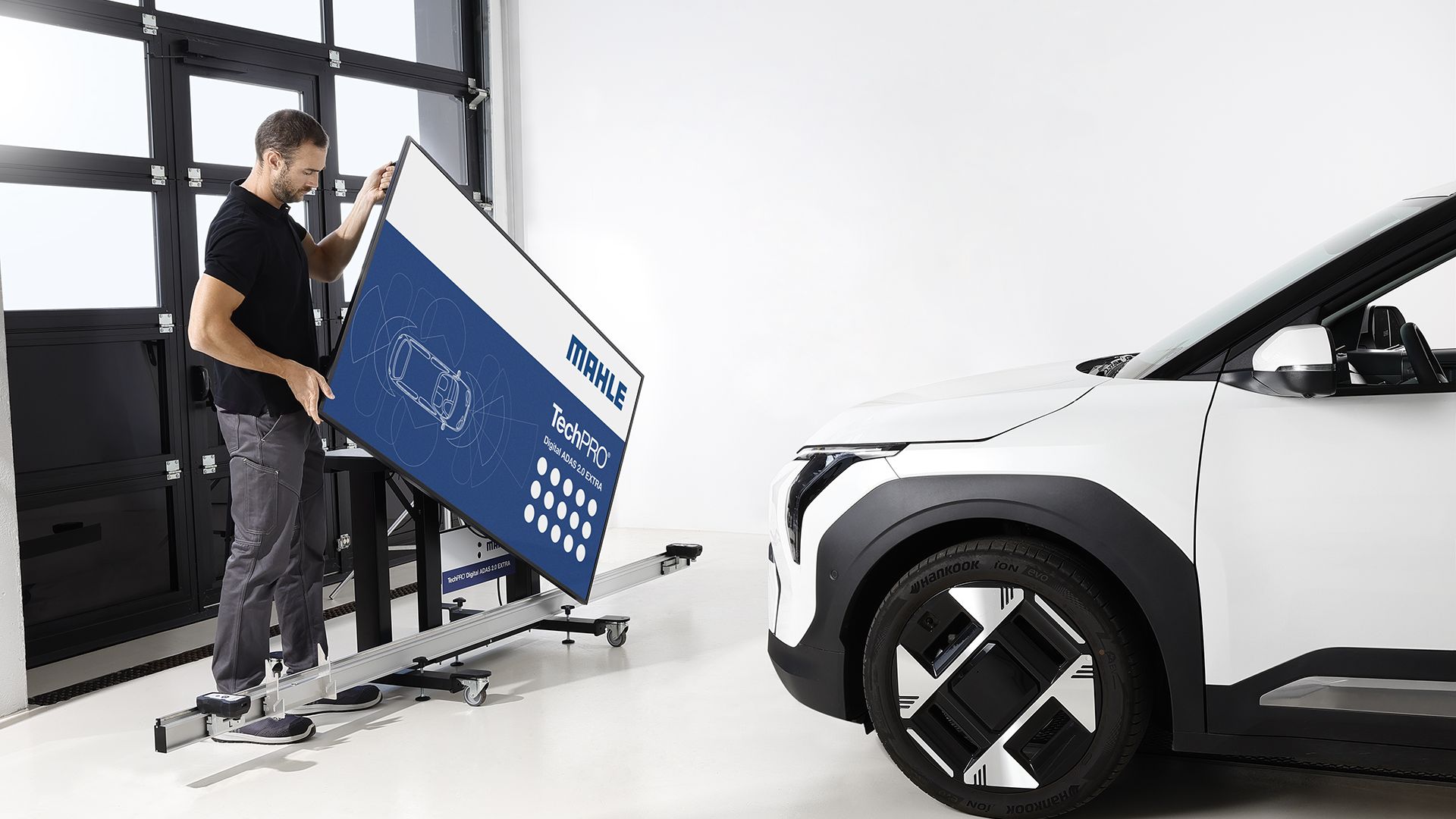 Un meccanico monta un grande pannello digitale per la calibrazione dei sistemi di assistenza alla guida MAHLE TechPRO Digital ADAS 2.0 EXTRA su una struttura mobile davanti a un veicolo bianco.