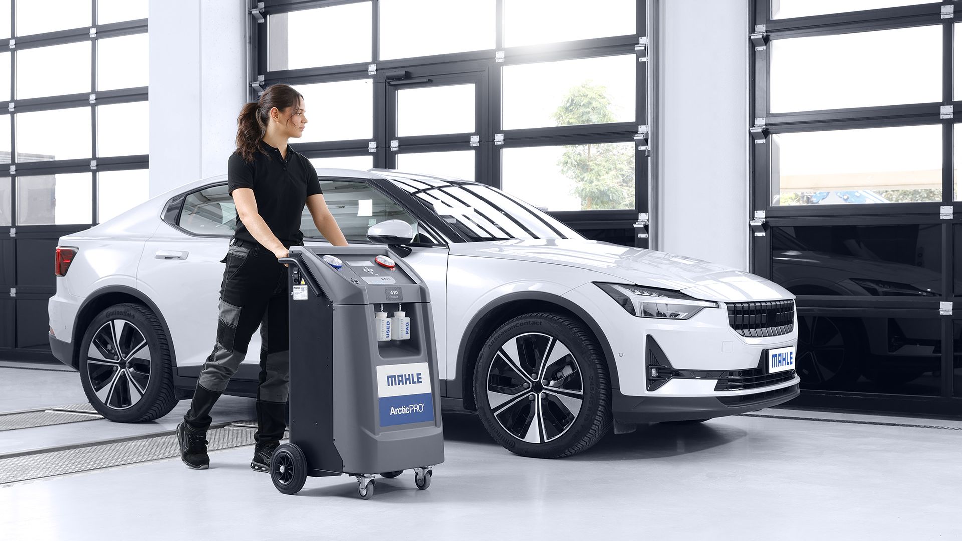 Una meccanica spinge una stazione di servizio per l'aria condizionata MAHLE ArcticPRO all'interno di un'officina, posizionata davanti a un'auto bianca.